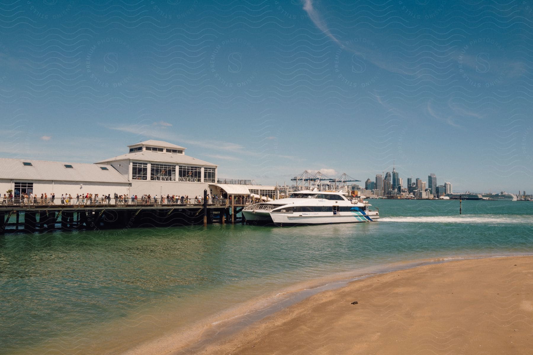 People stand on a boardwalk outside of a building overlooking the water as a boat sails with the metropolitan skyline in the distance