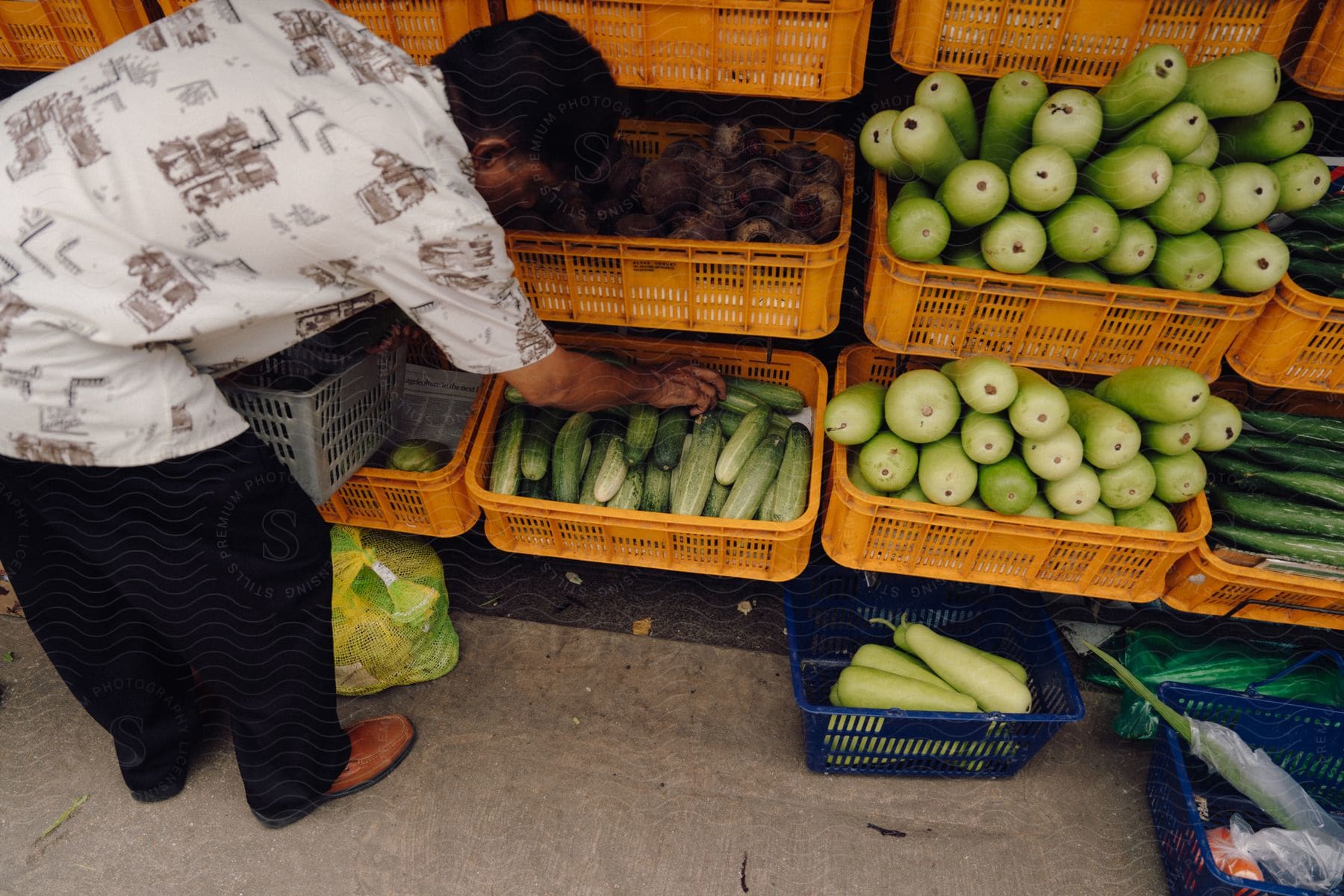 man wearing a white shirt and black pants takes cucumbers from a box at the fruit display shelf.