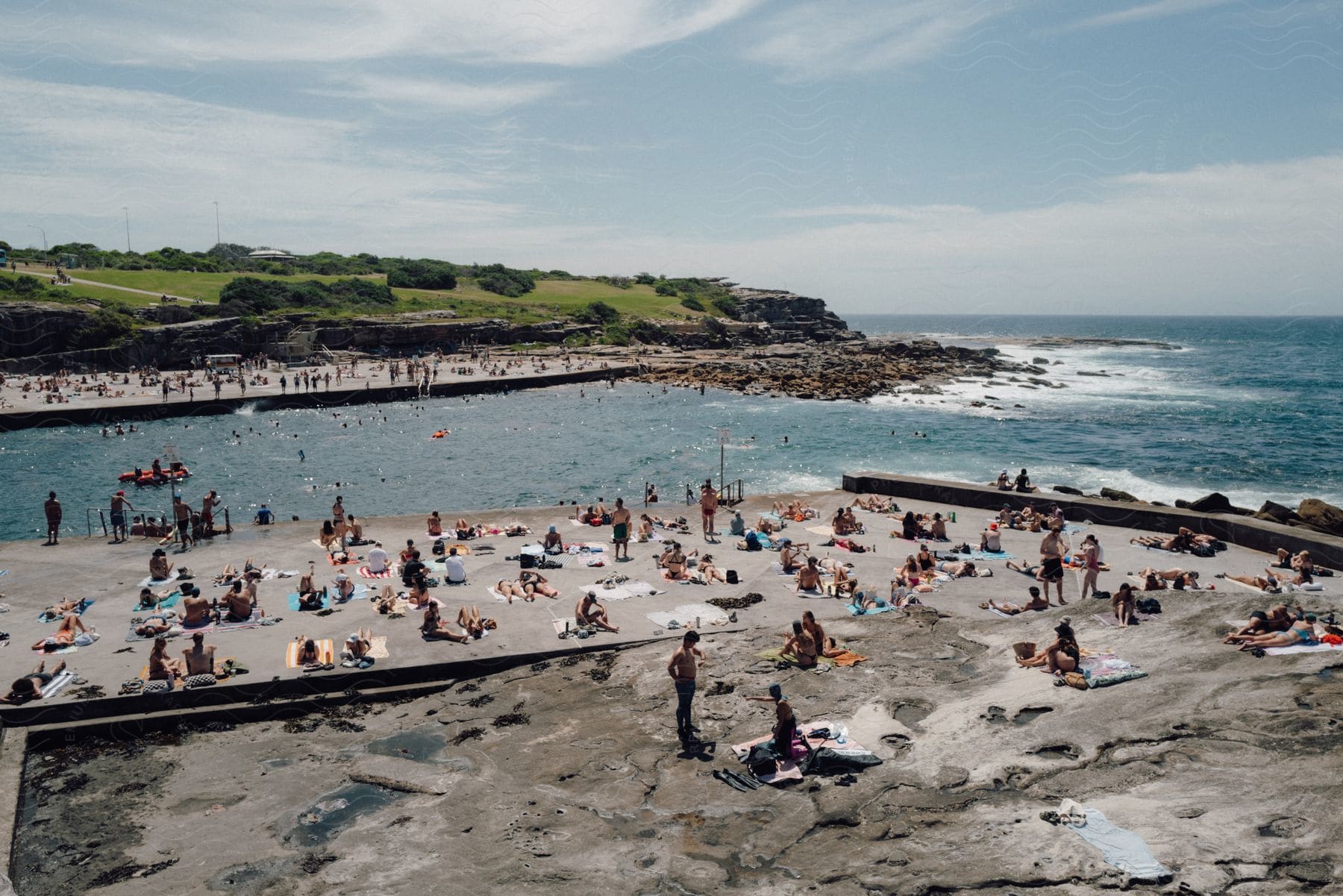A busy scene on a rocky beach, with people sunbathing and swimming during the sunny day.