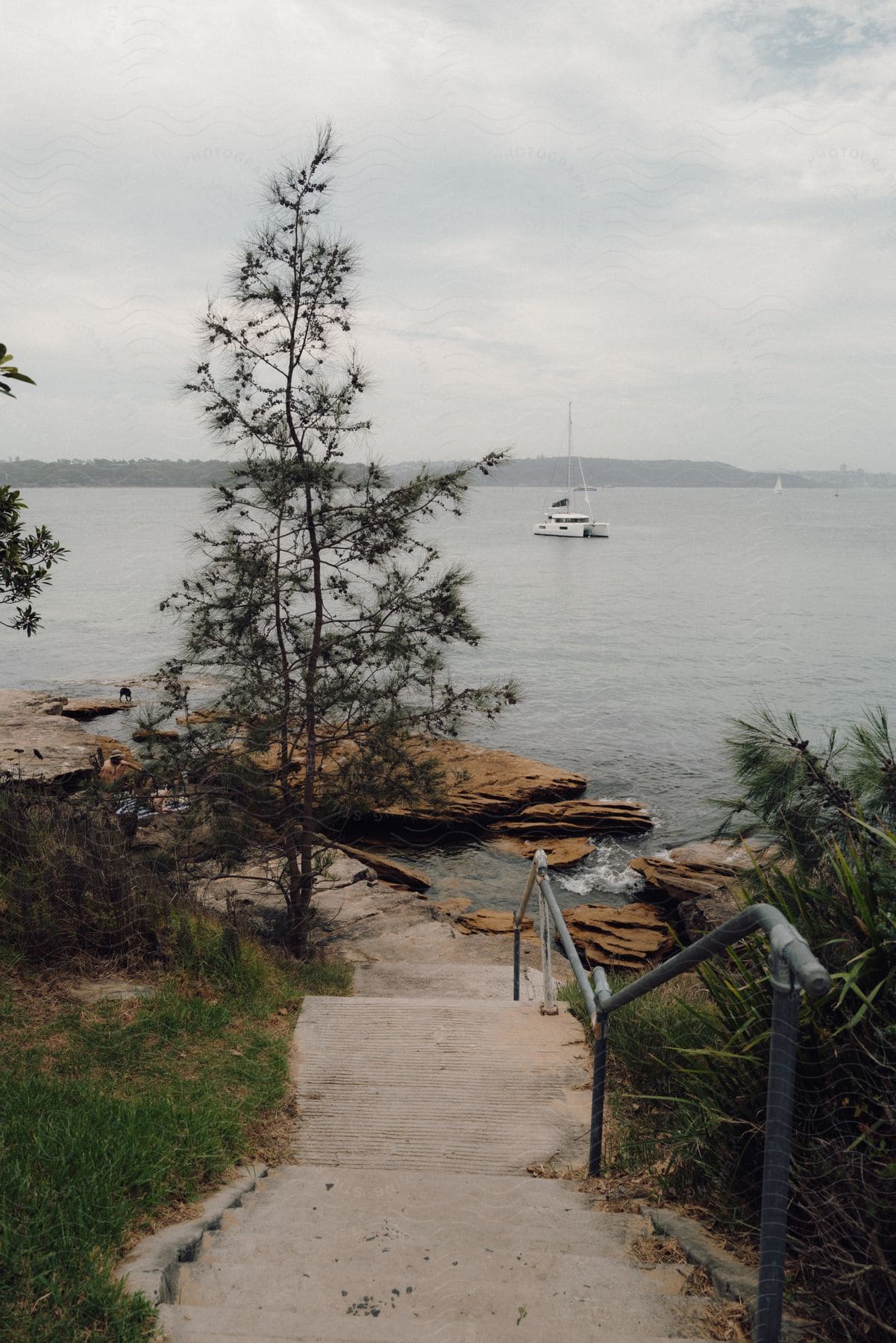 A serene view of a path leading to a rocky shore, with a yacht anchored in the calm waters.