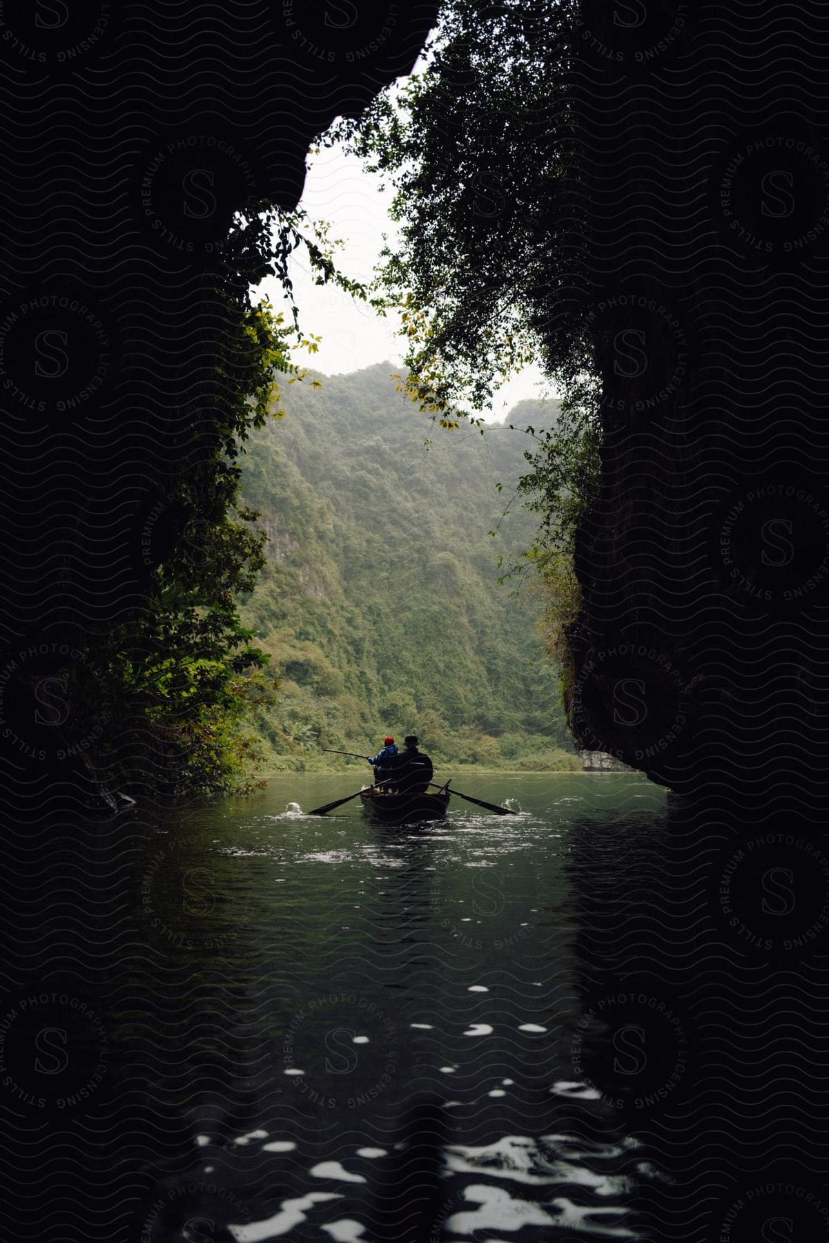 Two people paddle canoe through cave near mountain.