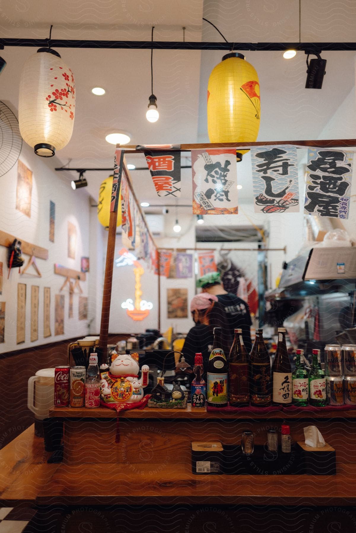 Interior of a Japanese restaurant with traditional decor including hanging paper lanterns and colorful flags. Two people are working behind the counter, surrounded by a variety of drinks.