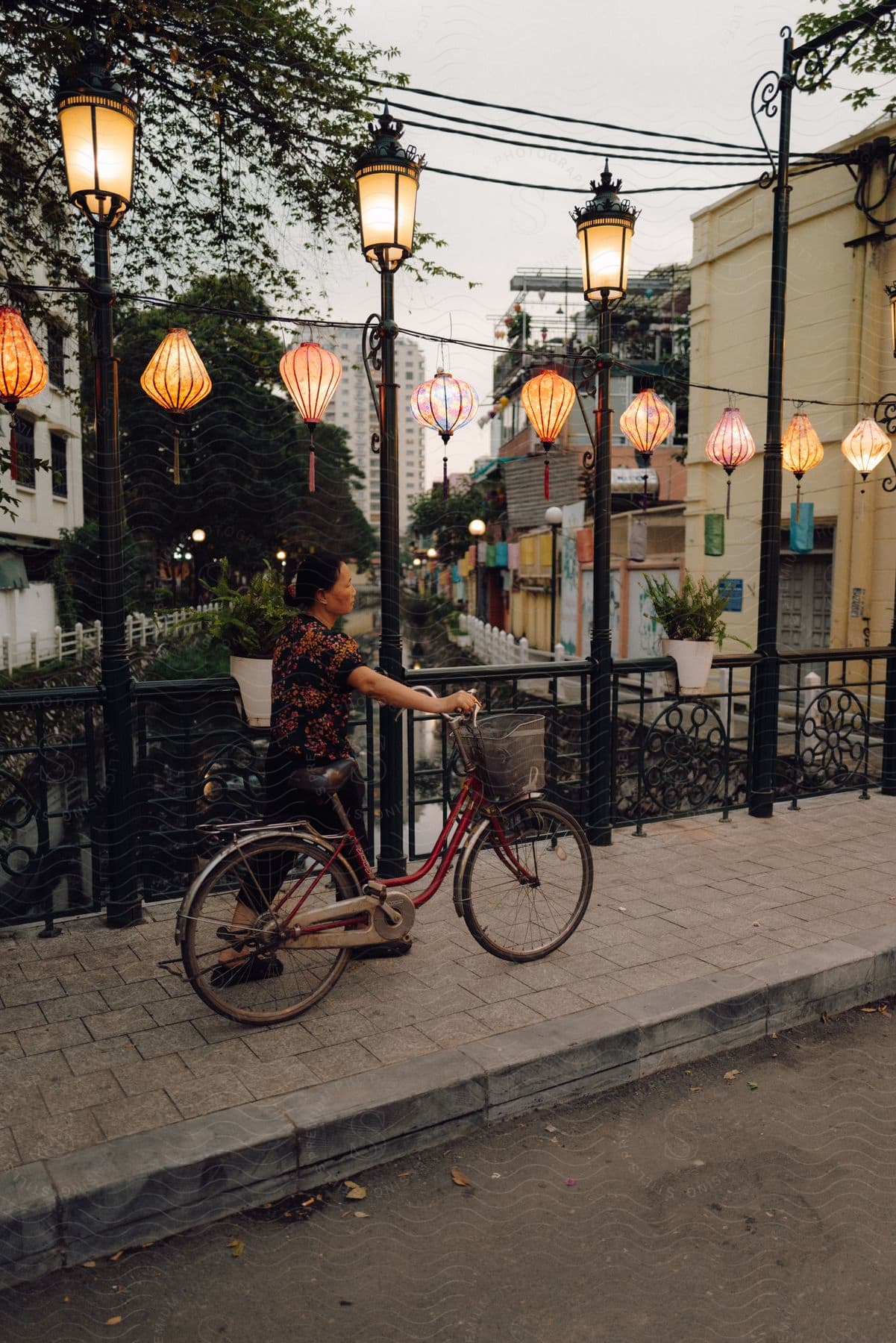 A woman walks her bicycle on a sidewalk under hanging lanterns, with buildings in the background.