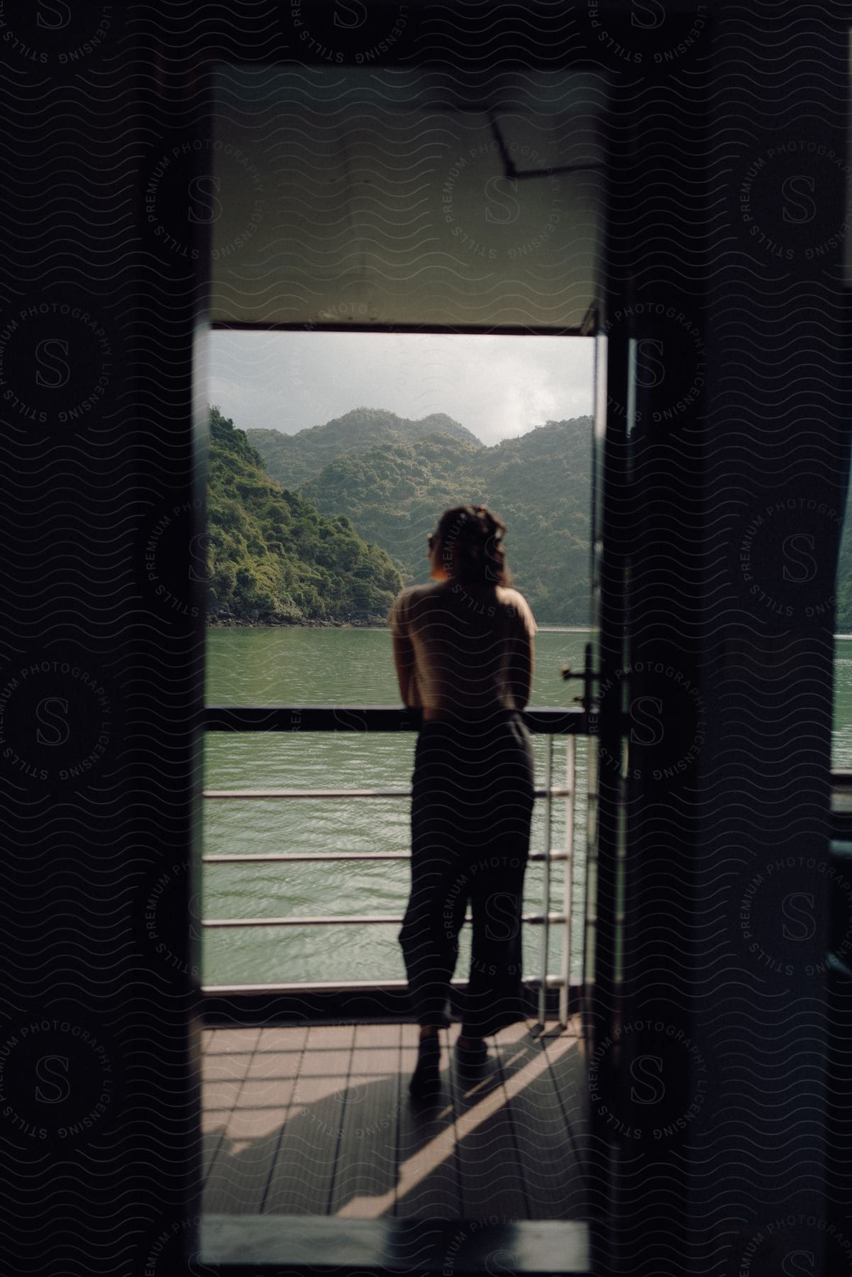 Woman stands on a balcony overlooking a green mountain landscape across the water.