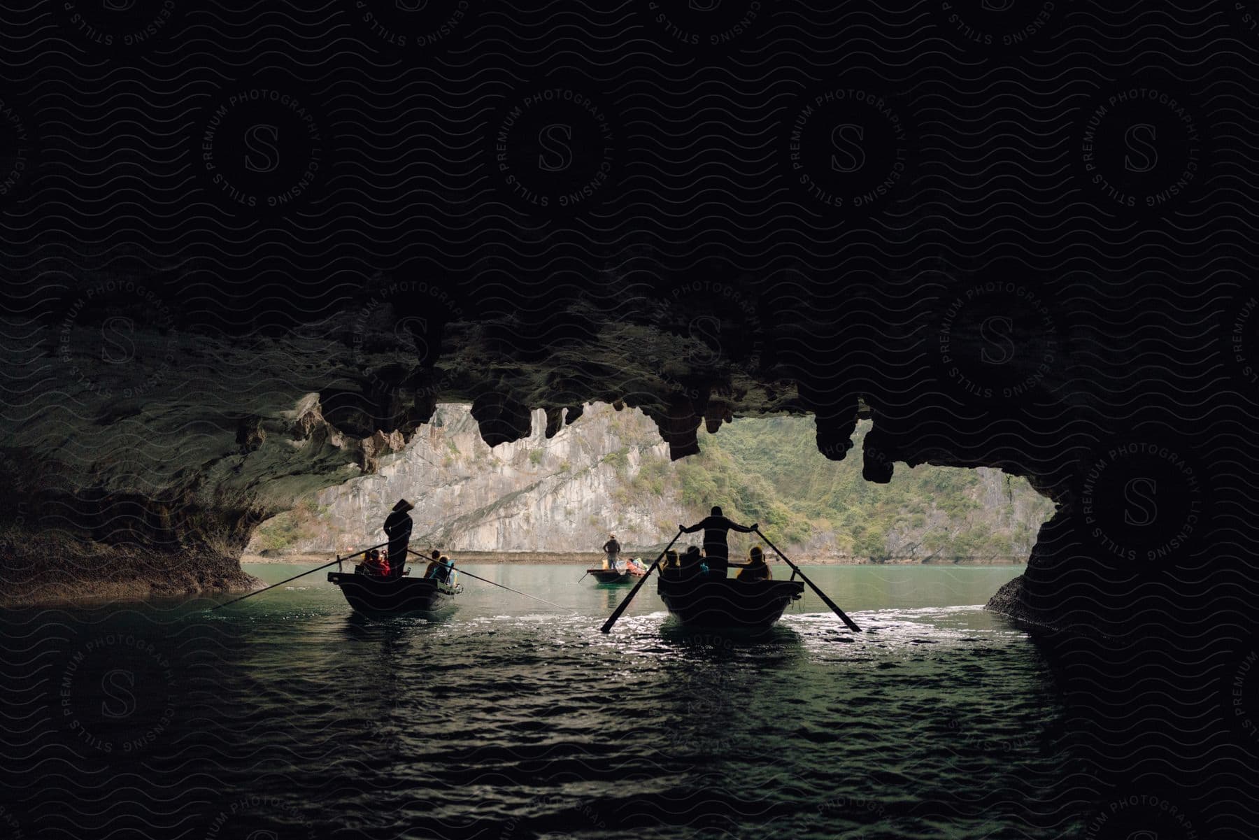 Silhouetted boats rowed by figures, navigating through a dark, cavernous waterway with rocky overhead formations, illuminated by natural light at the entrance.