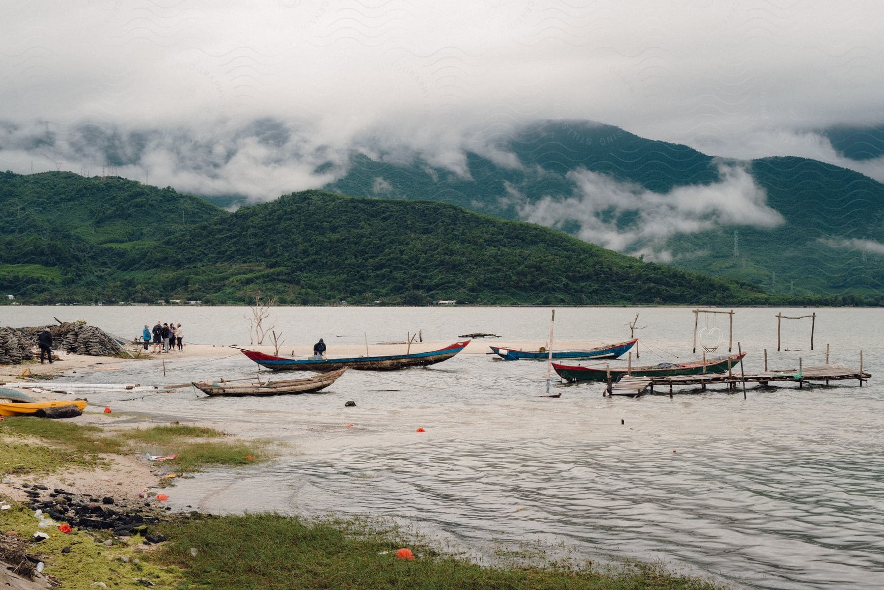 Boats moored on the coast across a bay from forested mountains.