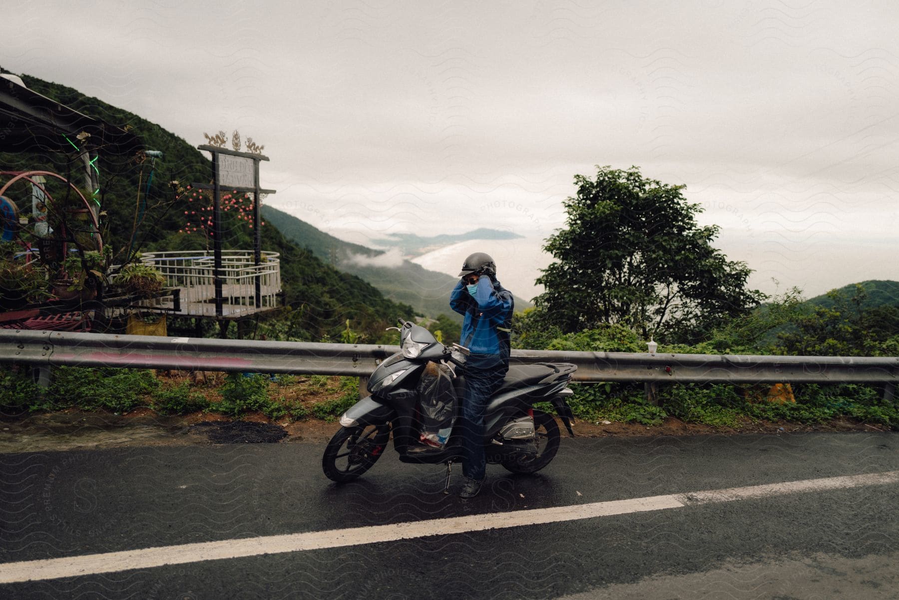 A person is putting on his helmet while sitting on a motorbike that is parked on the shoulder of an asphalt street