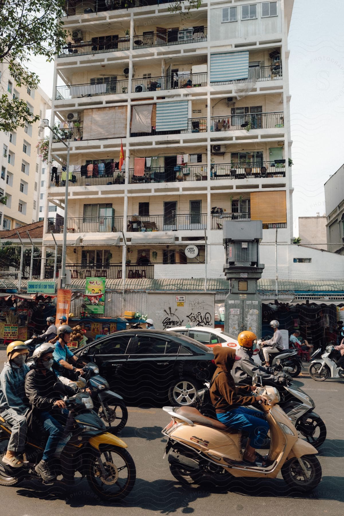 Several people riding motorbikes on a street and behind there are some cars and there is a huge building with clothes hanging on balconies in the background
