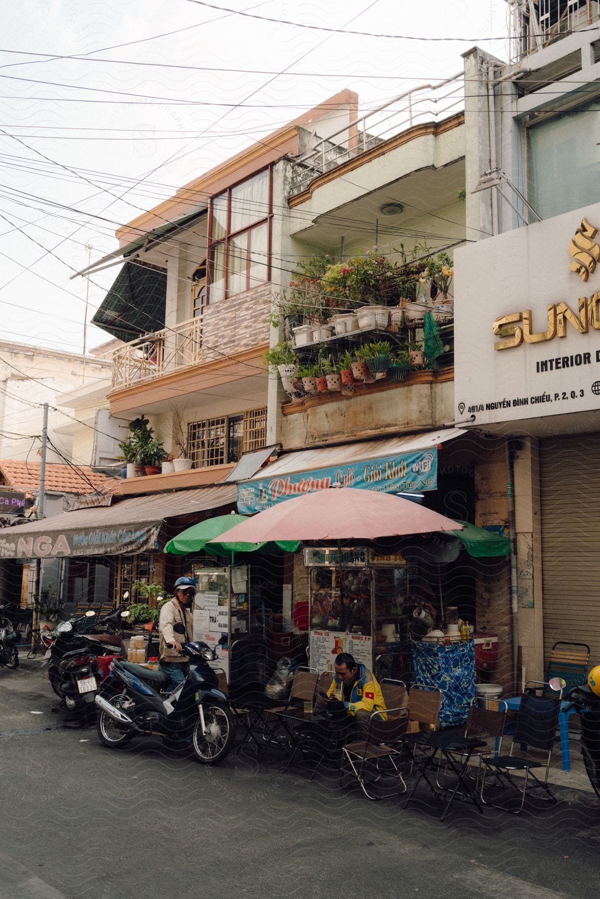 A street with some businesses on the sidewalk and some motorcycles on the street, there are two men, one standing and the other sitting on a chair