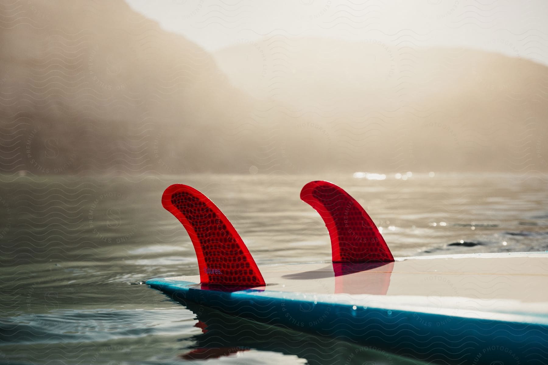 Close-up of red surfboard fins on calm water with hazy sunlight and mountain silhouette in the background.