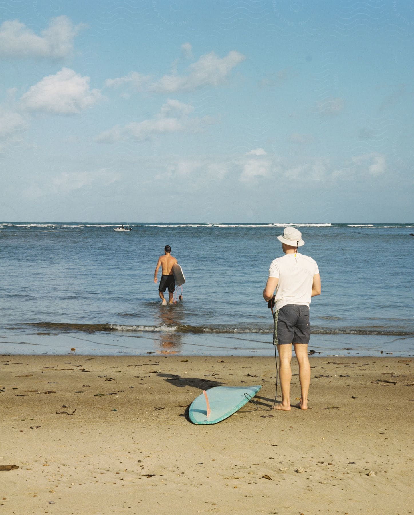 Man standing on the beach near his surfboard as he watches another man walking into the water with his surfboard under his arm