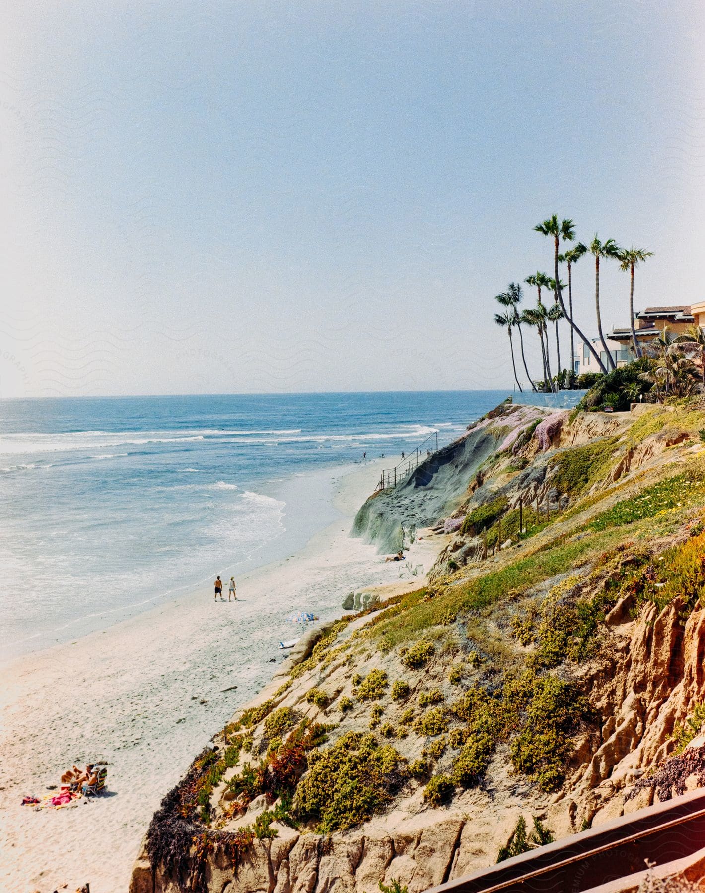 Sandy beach with a few people, towering palm trees on the cliffside, and a clear blue sky above.