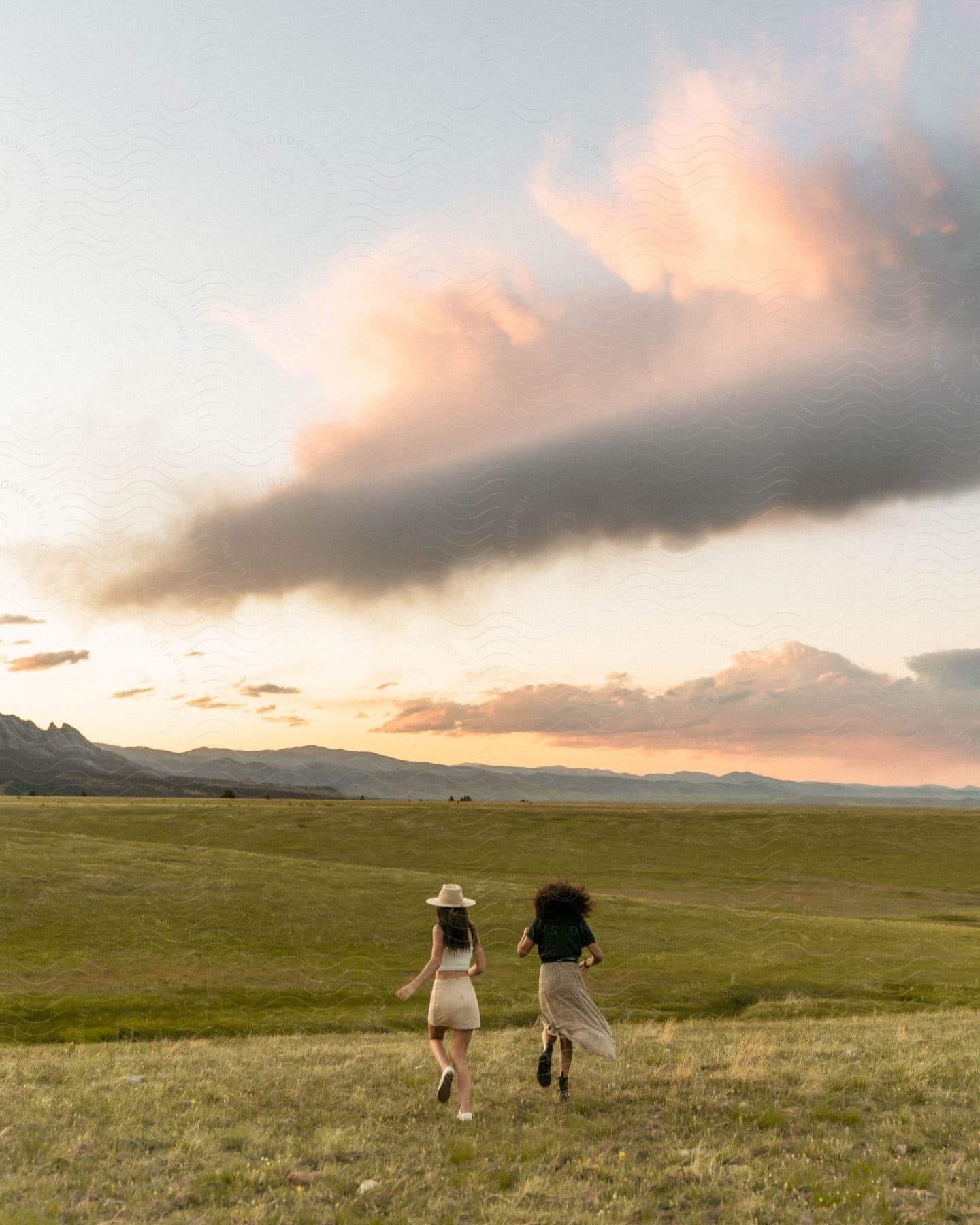 Two women, one wearing a hat, a white blouse, and shorts, and the other wearing a black shirt and a skirt, joyfully run down the green field