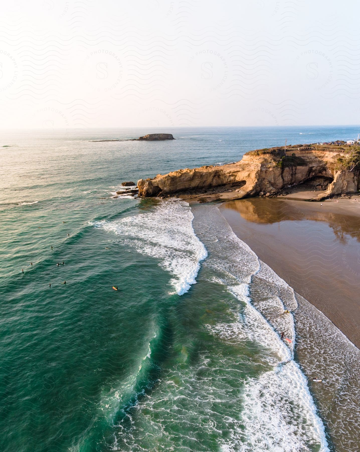 Aerial view of a beach with clear waters and waves gently breaking on the sand with the rocky coast in the background and surfers in the middle of the sea.