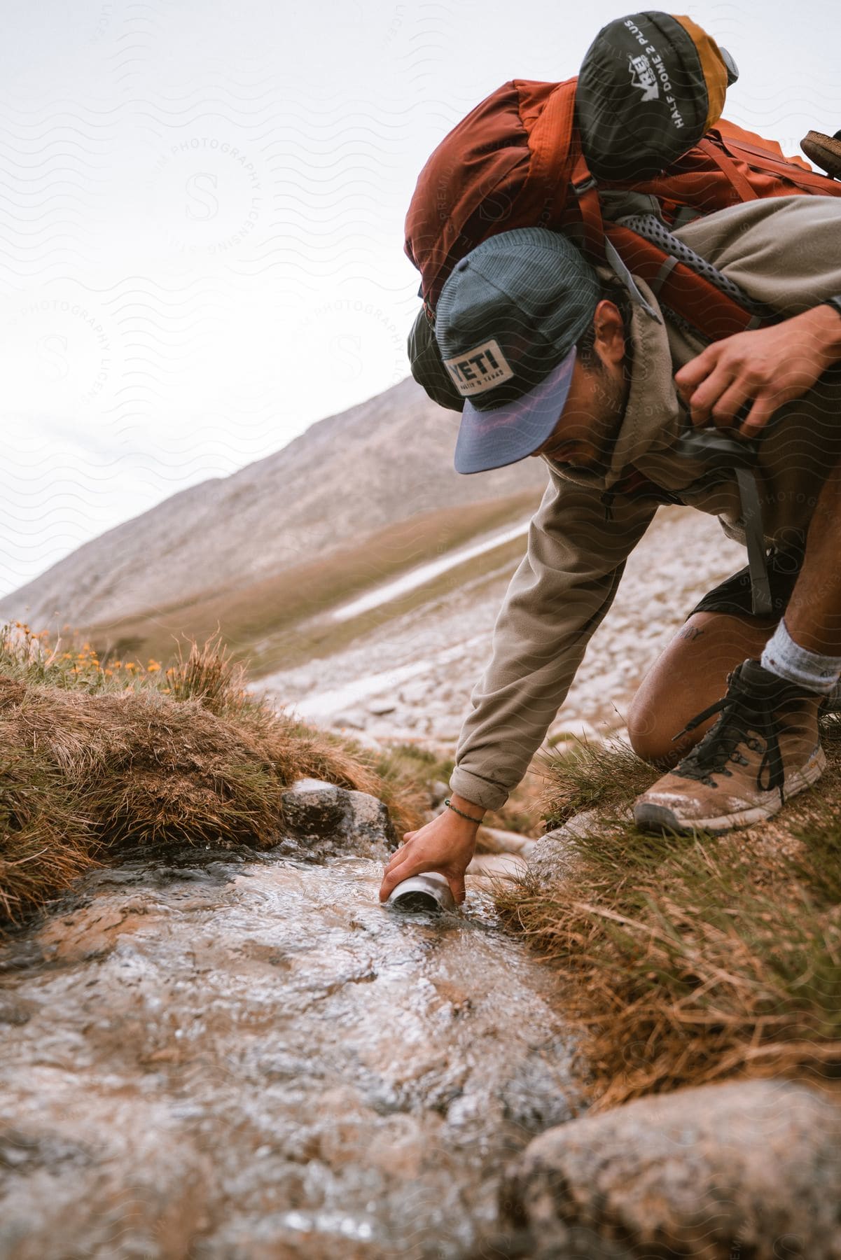 A traveler bending down to collect water from a stream in mountainous terrain.