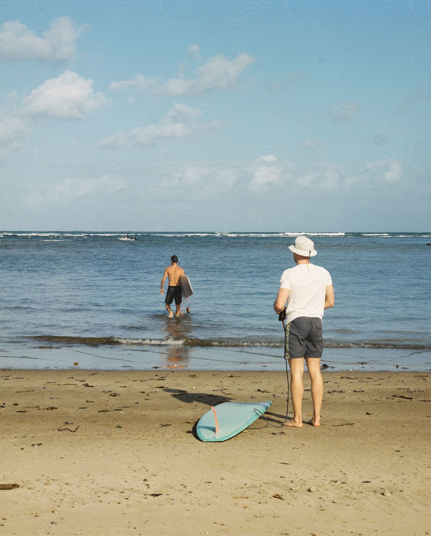 One man wades in the water with his surfboard while another watches from the beach.