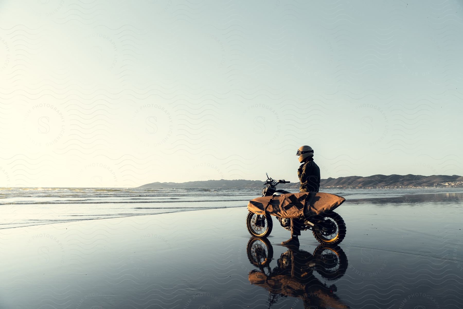 Person sits on a motorcycle with a surfboard on the coast near low mountains.