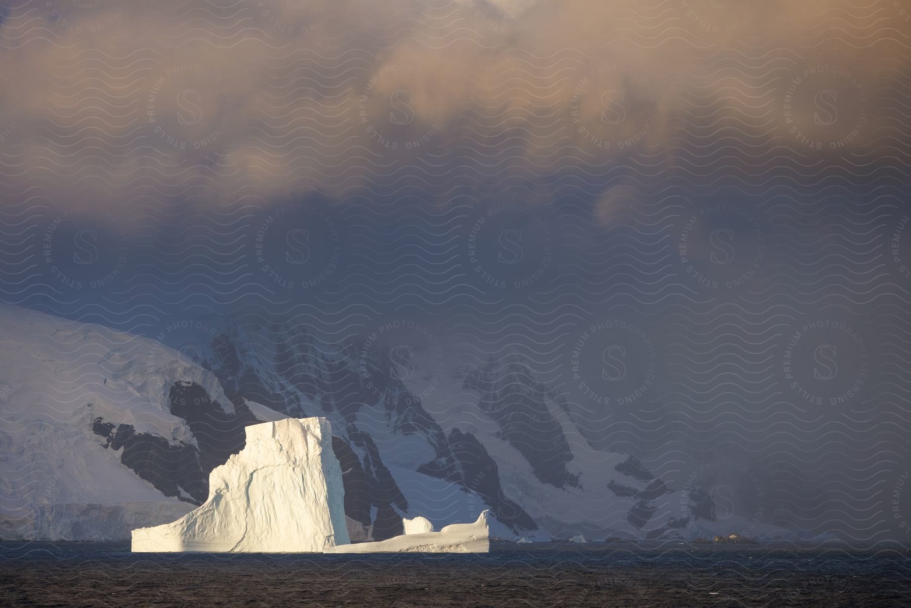 Iceberg floating in the ocean with snow-capped mountains in the background in a cloudy sky.