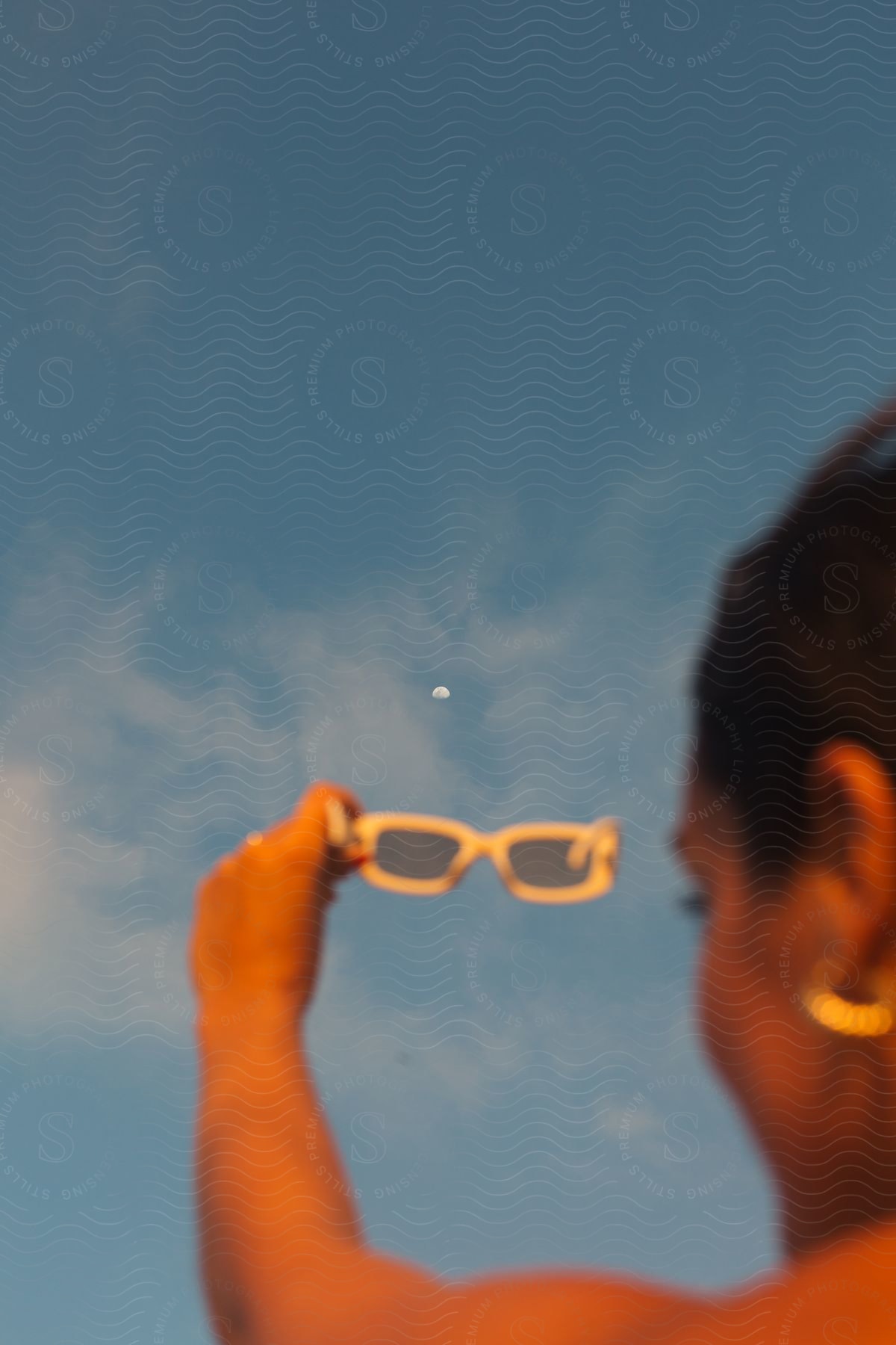 A person holds a pair of sunglasses against a blue sky with a visible moon.
