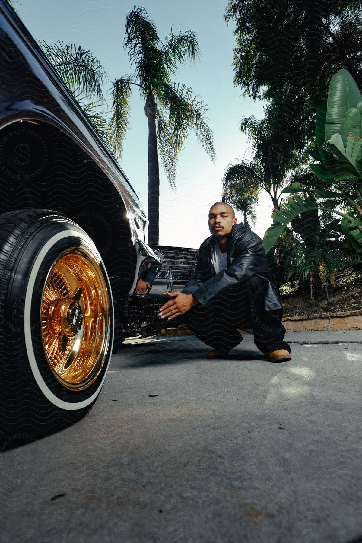 Man in leather jacket crouching posing next to two vintage vehicles with chrome wheels