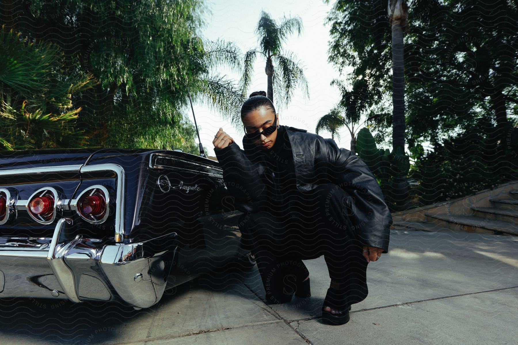 Female wearing leather jacket and sunglasses crouching next to a car posing outdoors