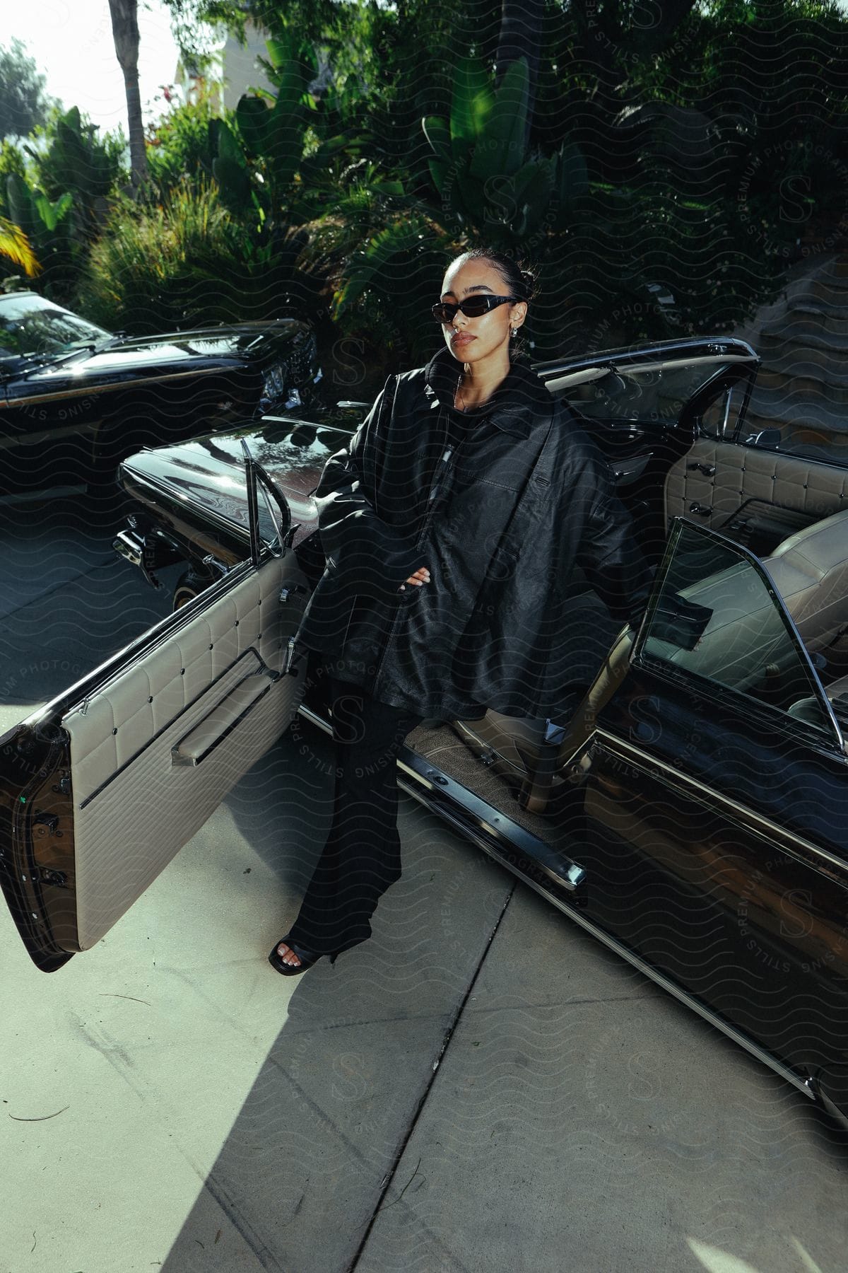 Woman posing next to a classic car with the door open and wearing a black leather outfit and sunglasses