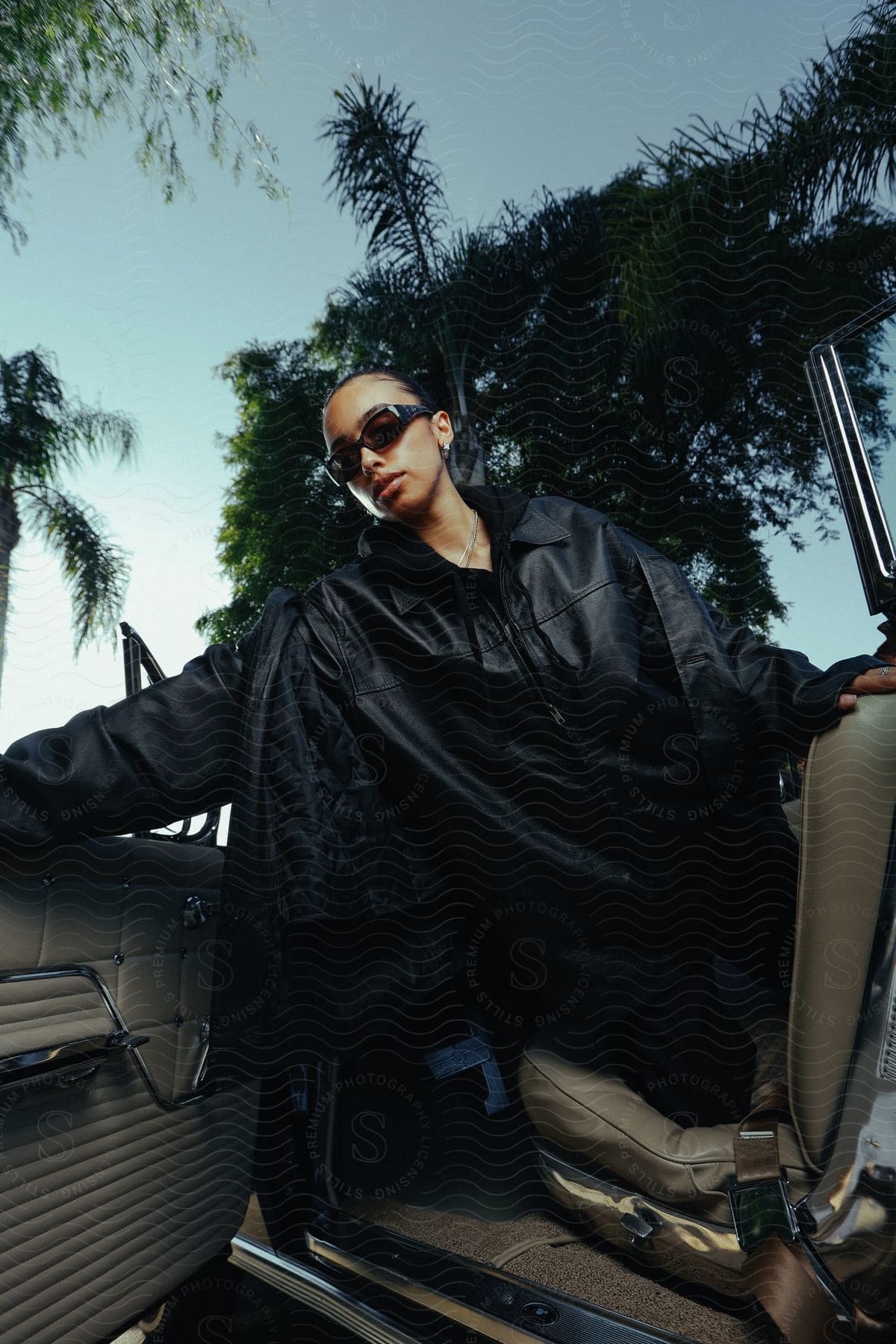 A young woman leans out the driver's side door of a convertible while modeling an all black outfit.