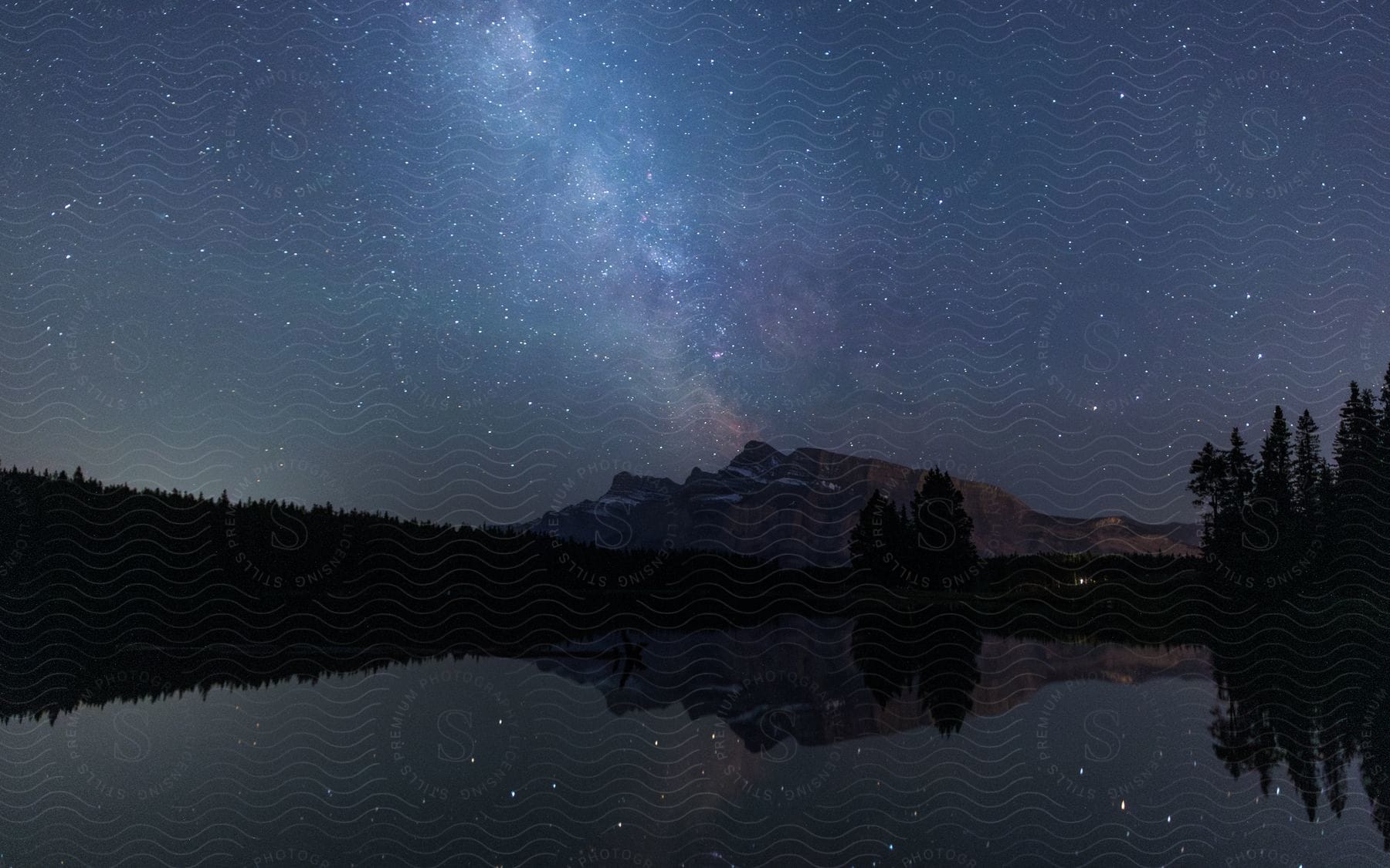 Starry Night Sky With The Milky Way Over A Silhouette Of Mountain Peaks And A Calm Lake Reflecting The Stars