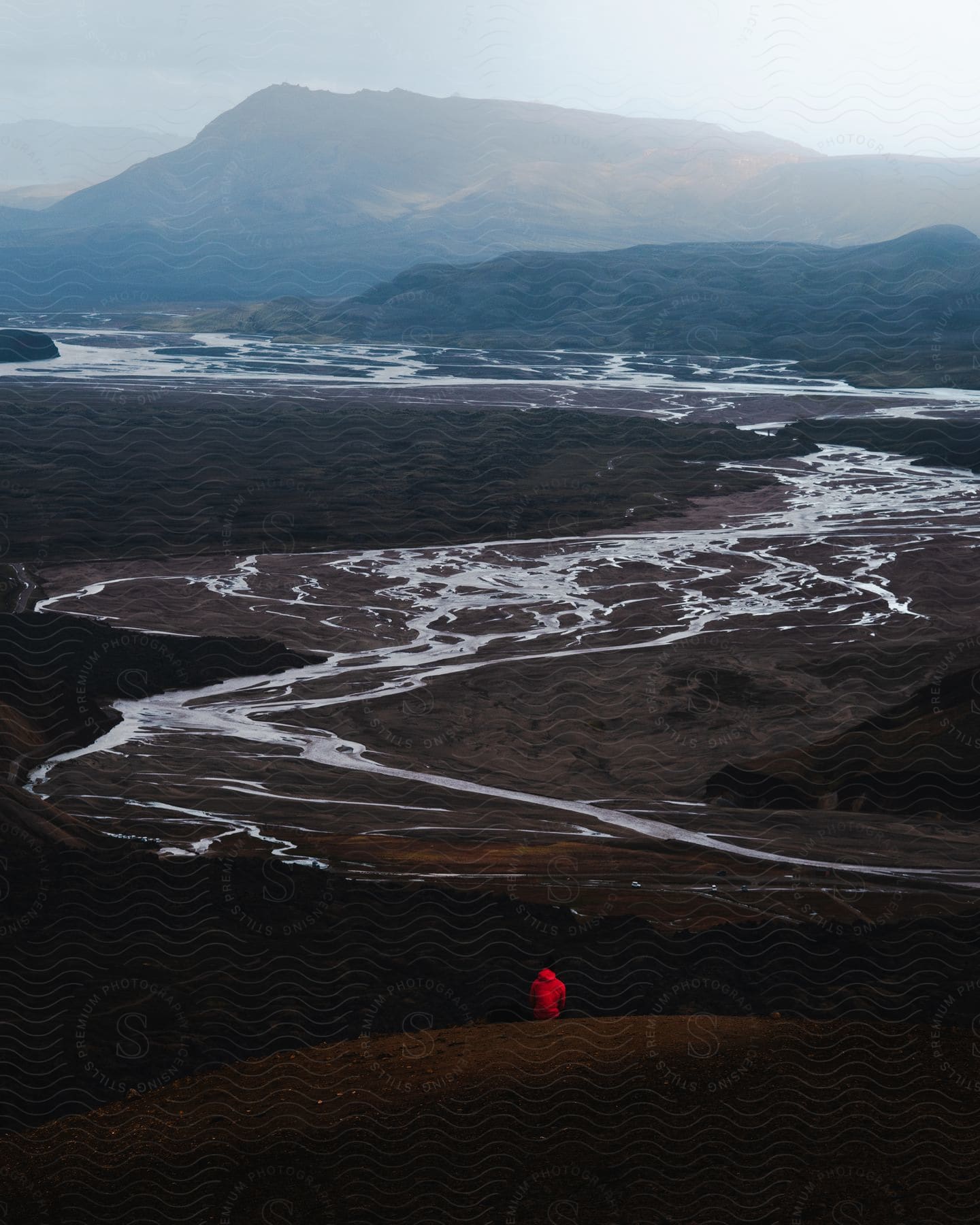 A person in a red jacket is sitting on a hill and watching a river that is dry