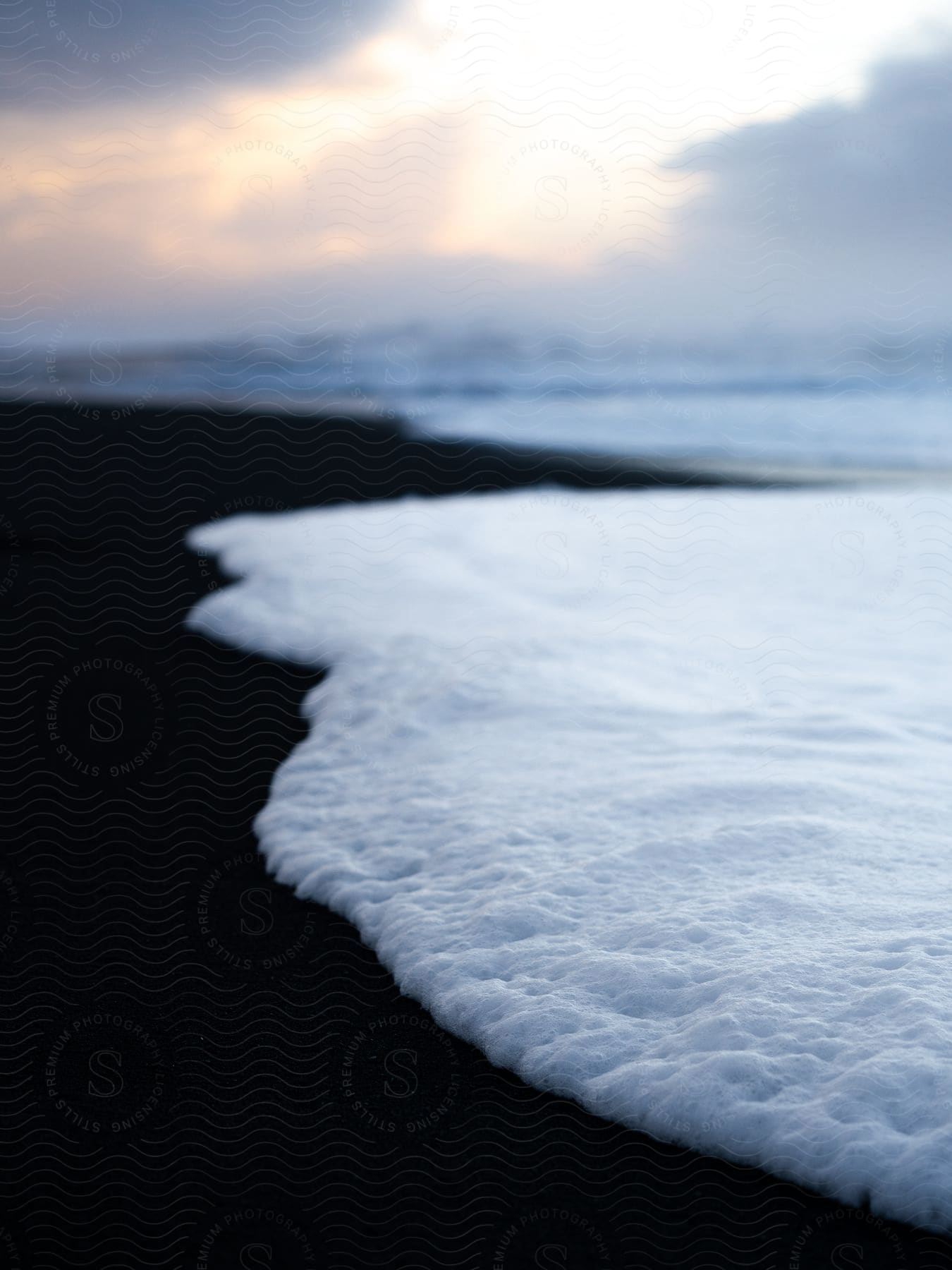 A foamy ocean wave breaks against the black sand beach at sunset.