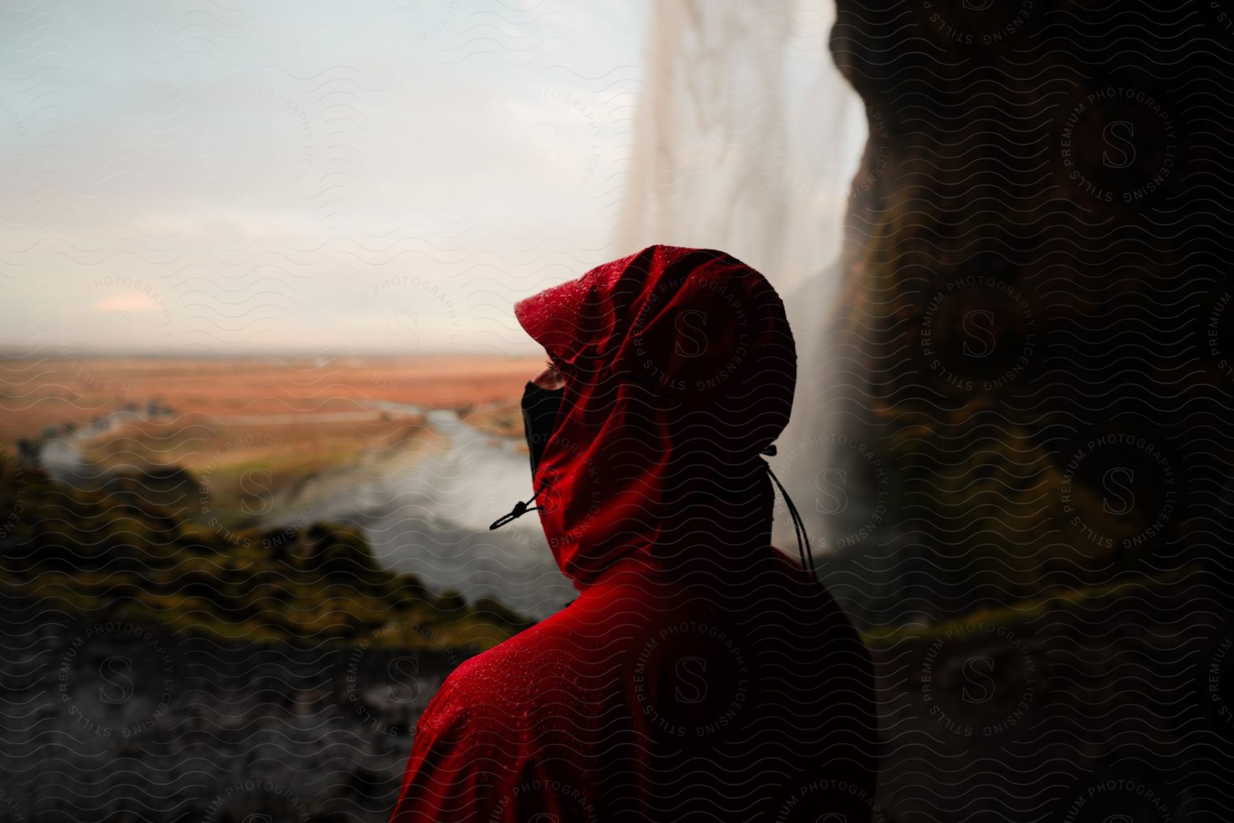 A person with his back turned and wearing all red clothing and his face is covered with a mask and in front of him there is a waterfall coming out of a mountain