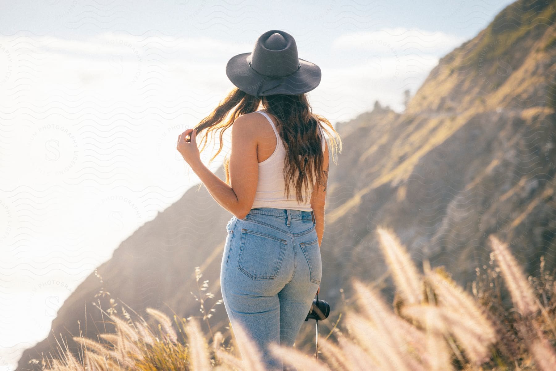 Woman wearing a sunhat plays with her hair as she stands looking at a mountain