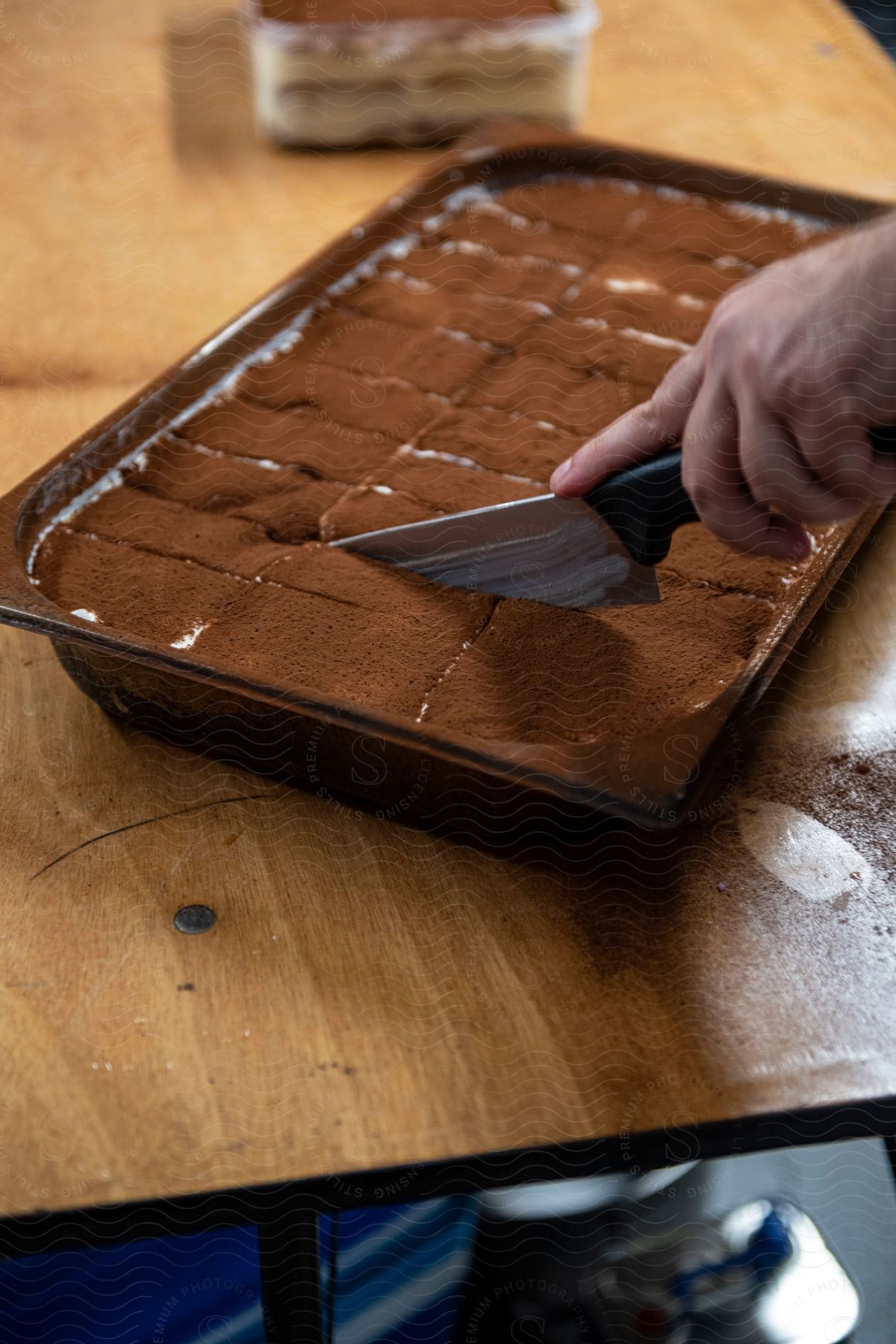 A human hand using a knife to cut a cake that is in a rectangular shape on a wooden table