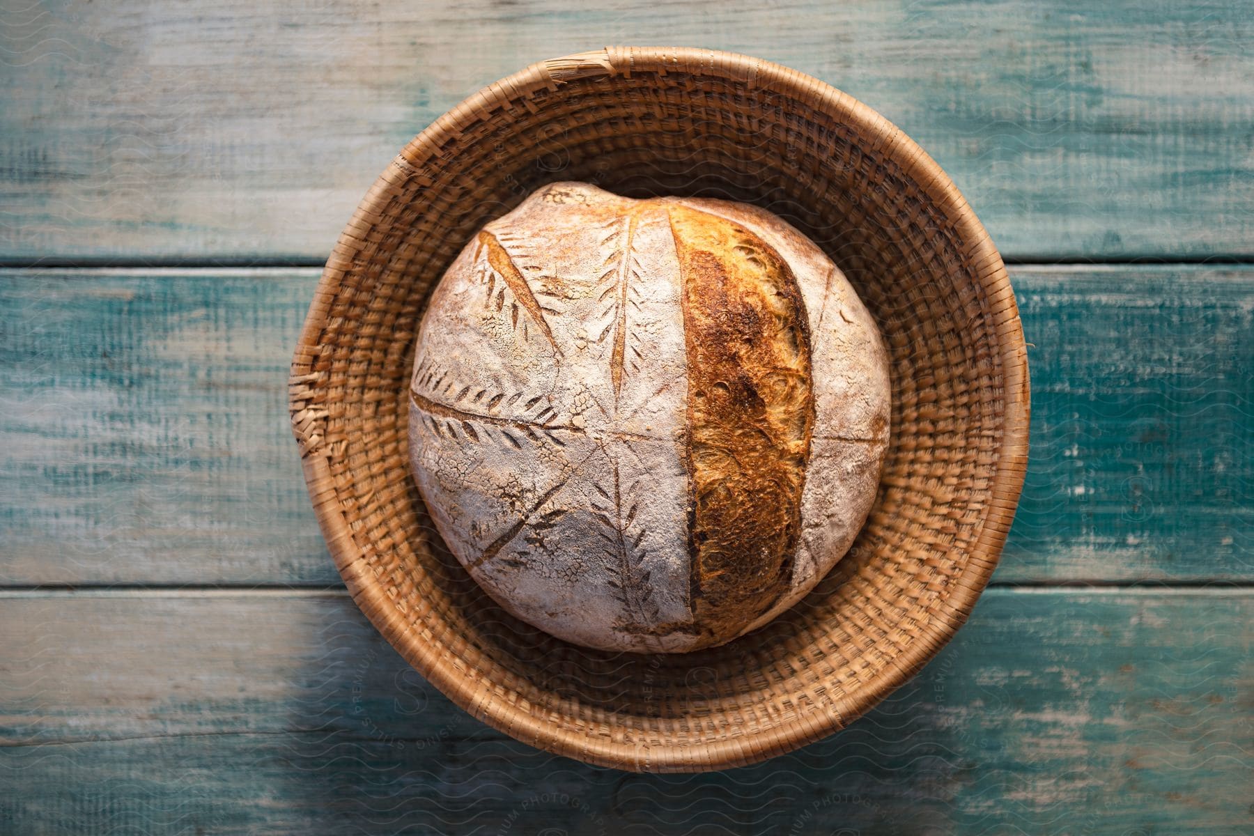 A wicker basket on a blue wooden surface contains a loaf of bread with leaf prints on the golden crust.