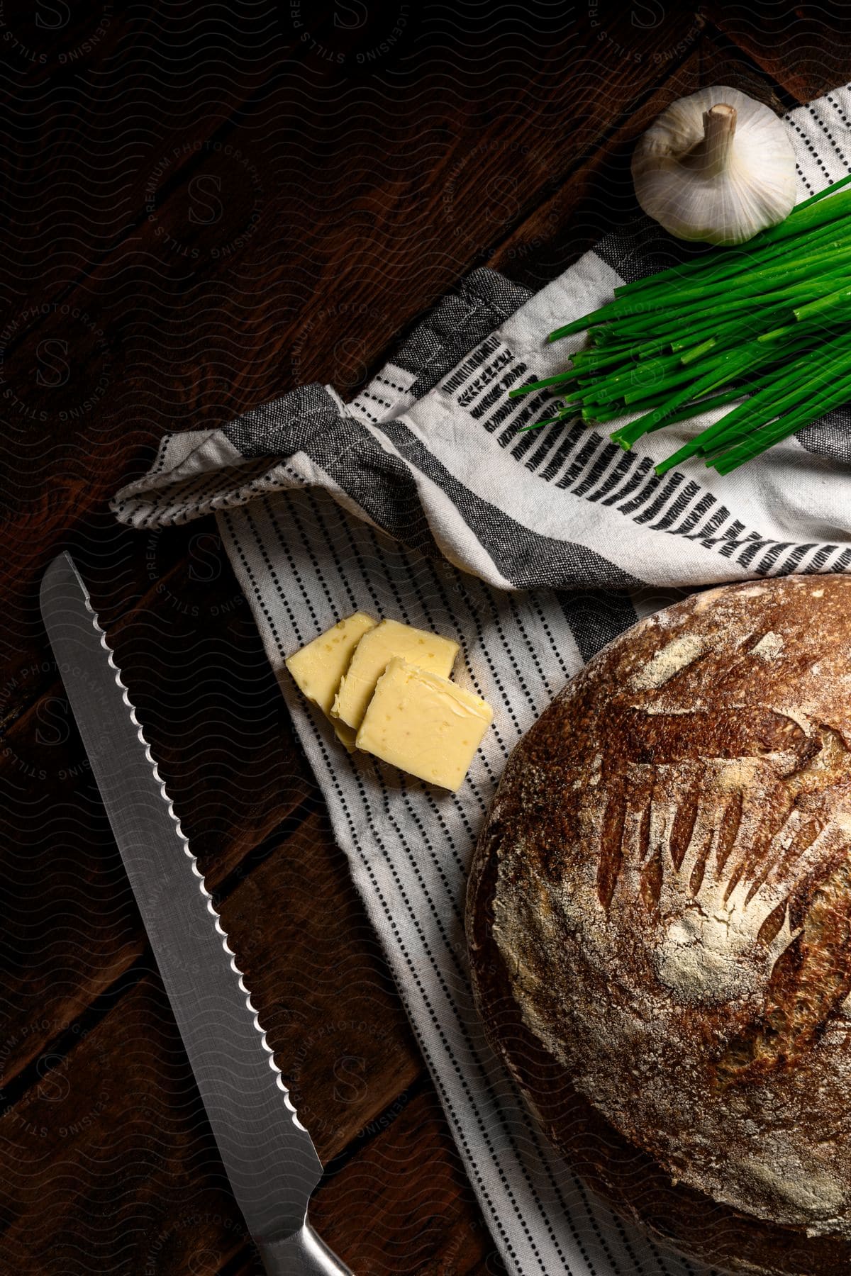 A loaf of bread,butter slices,green onions, garlic and a serrated knife on a dish towel.