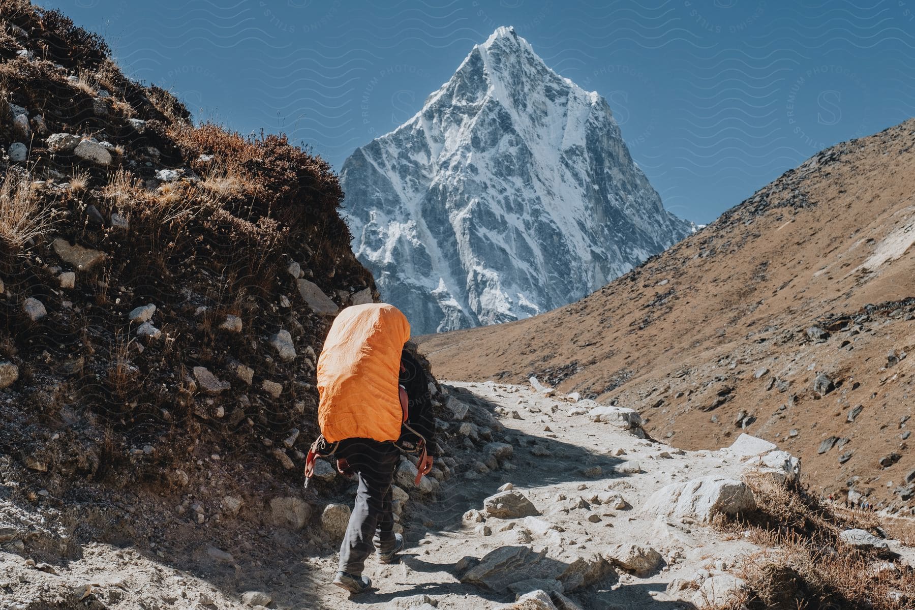 A hiker carrying an orange backpack walks up a mountain trail on a sunny day.