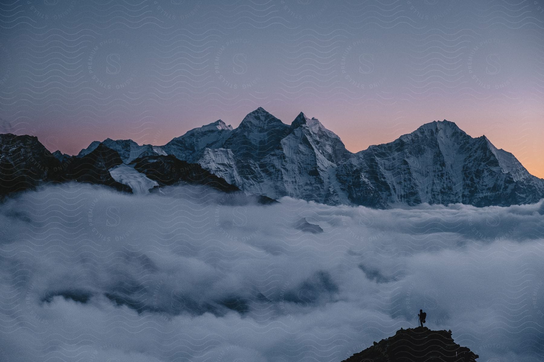 Stock photo of hiker standing on a ledge looking at a mountain range with rough peaks high in the clouds as the sun has set