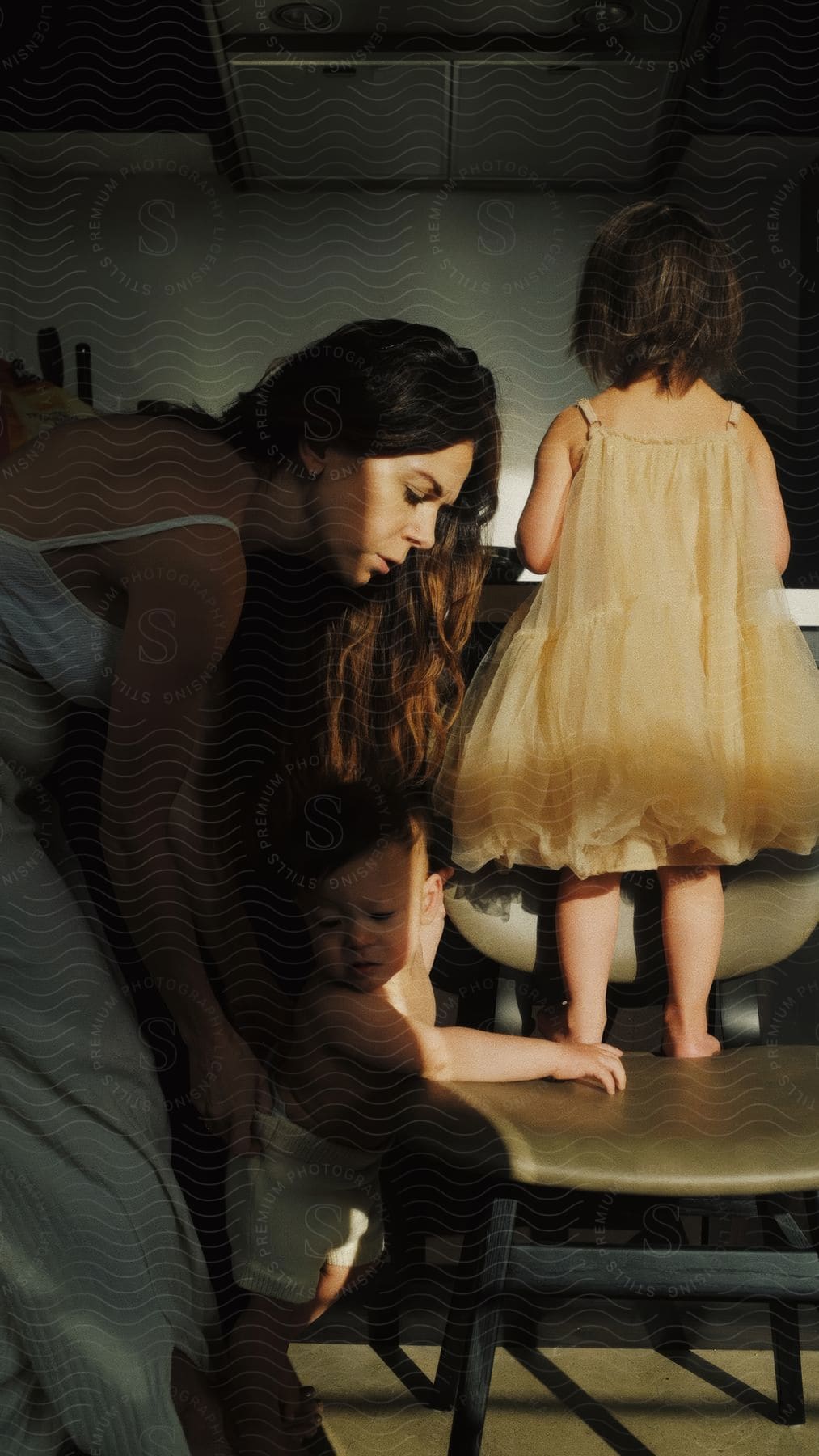 A woman in a kitchen holding a baby and a little girl in a yellow dress standing on a chair.