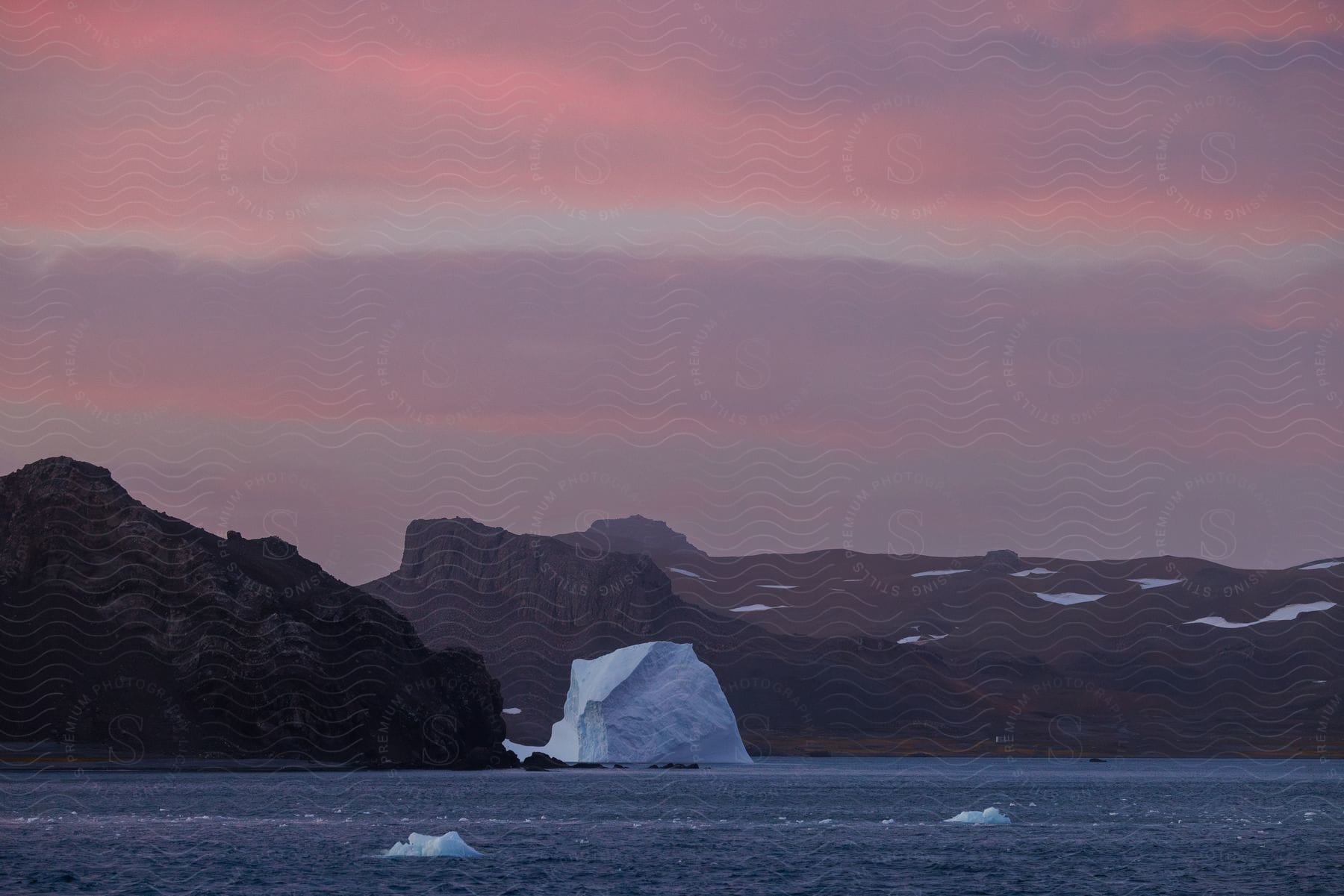 A completely white iceberg floating in the water and behind it there are some stone mountains with some snow