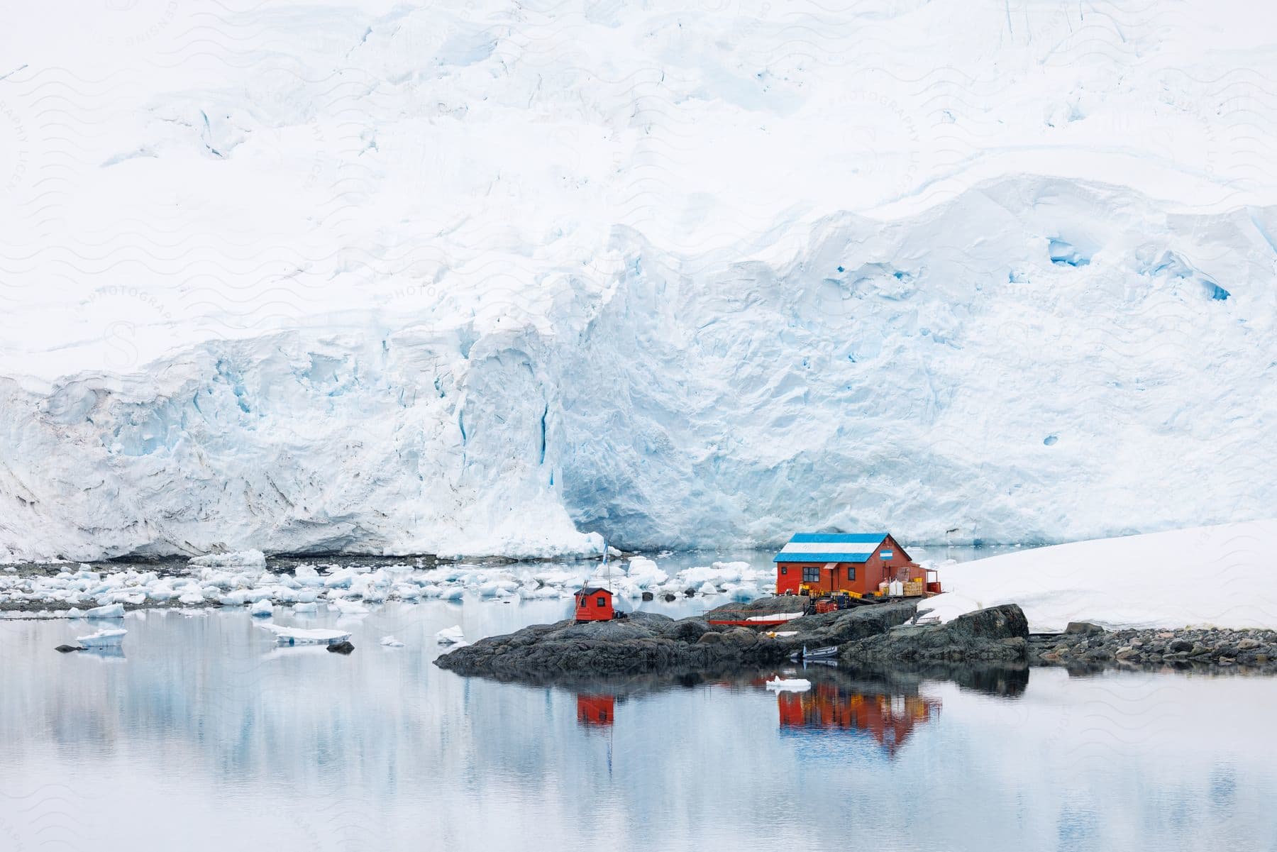 A small, weathered house with a red roof sits on a rocky outcropping, surrounded by a vast expanse of frozen ocean.