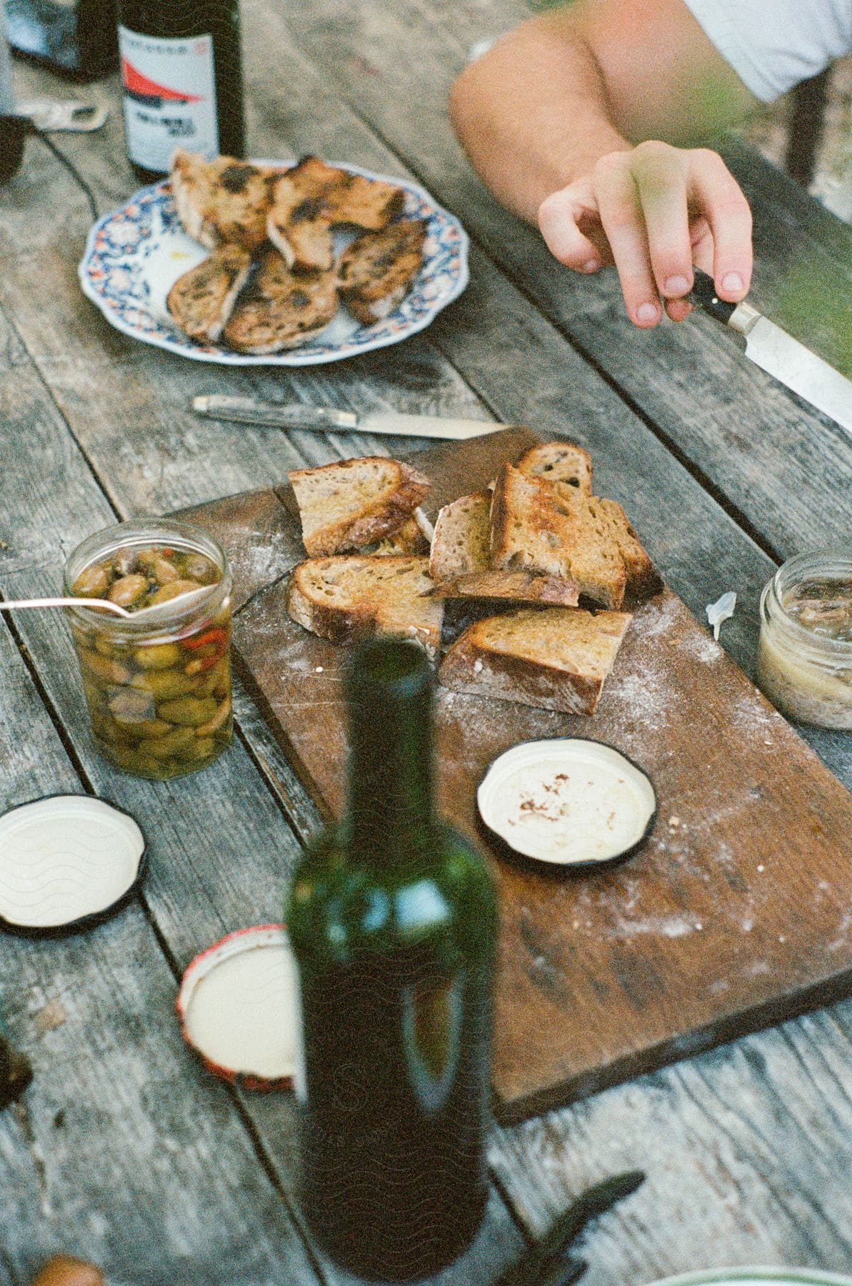 Rustic outdoor dining setup with toasted bread slices on a wooden cutting board, a jar of olives, and an open bottle of wine on a weathered wooden table.