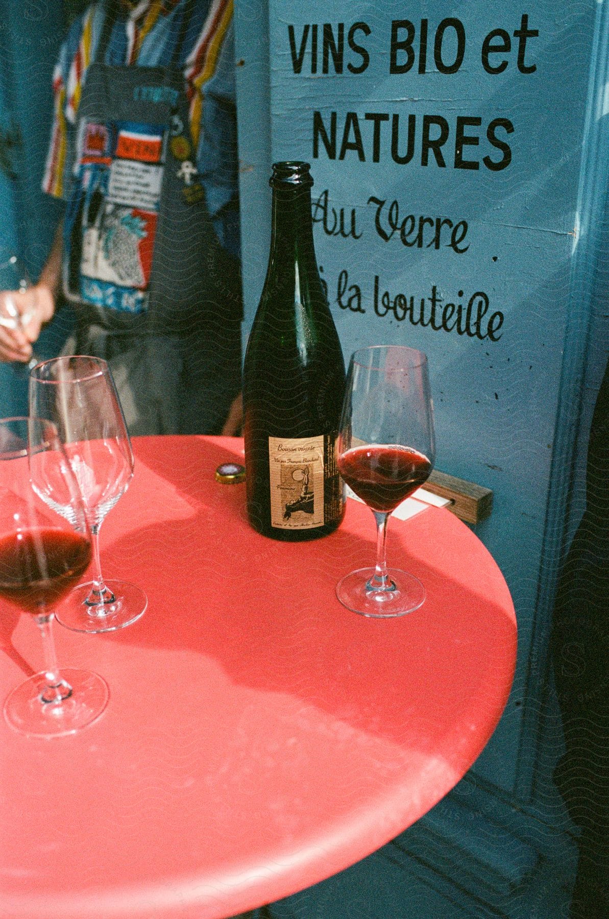 A bottle and two glasses of wine on a red table beside a blue board