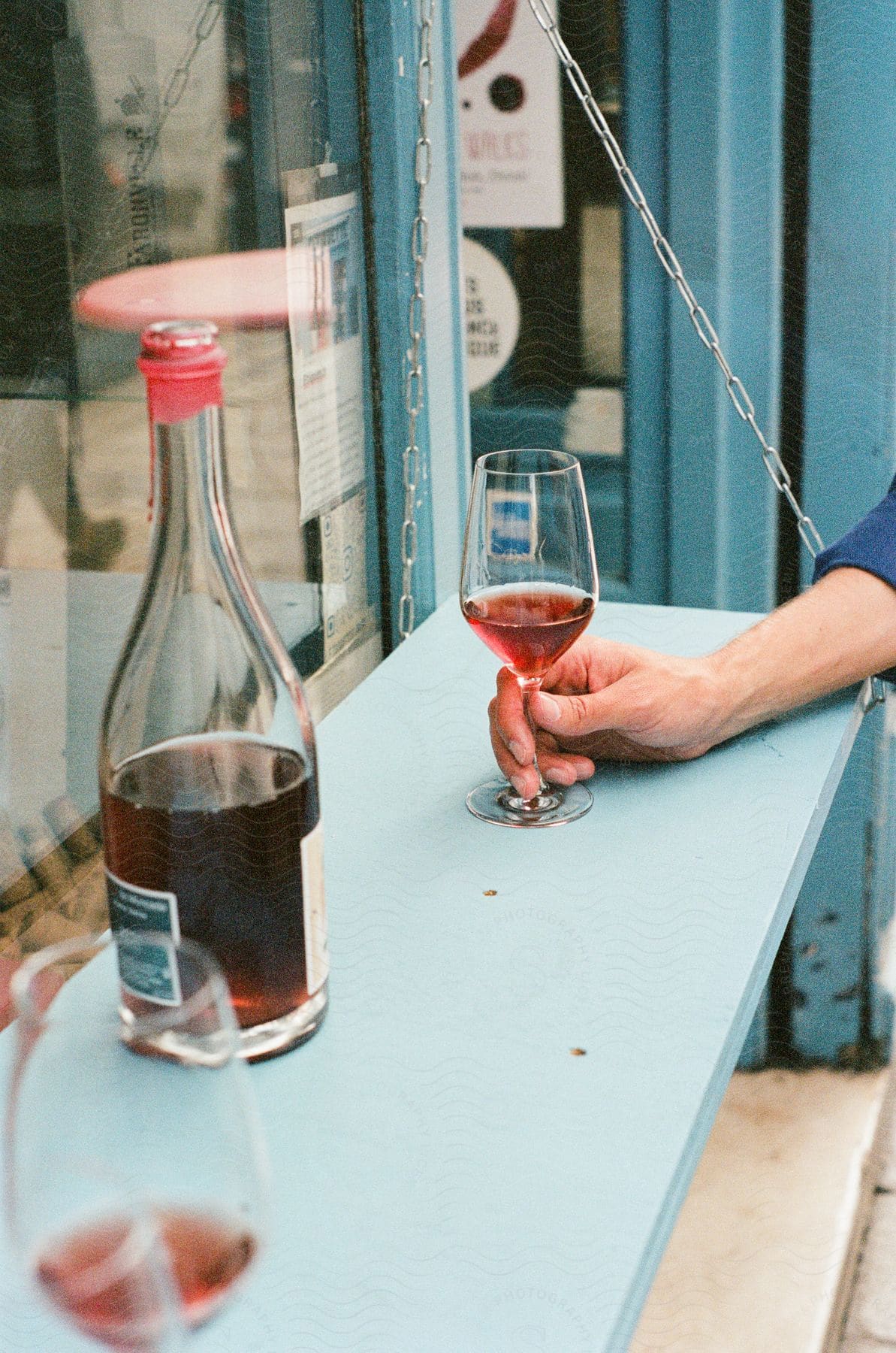 A man wearing a blue shirt holds a glass of wine next to a wine bottle on a window sill of the cafe.