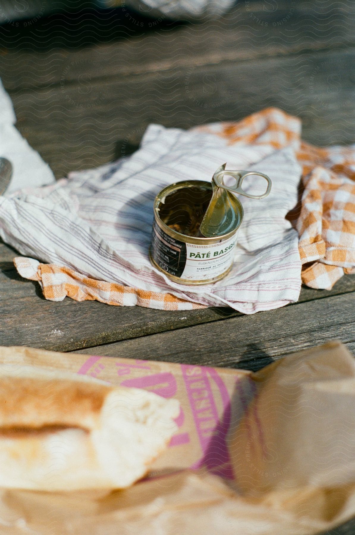 An open can of pâté on top of a cloth that is on a wooden table and in front of a package of bread with a loaf on top