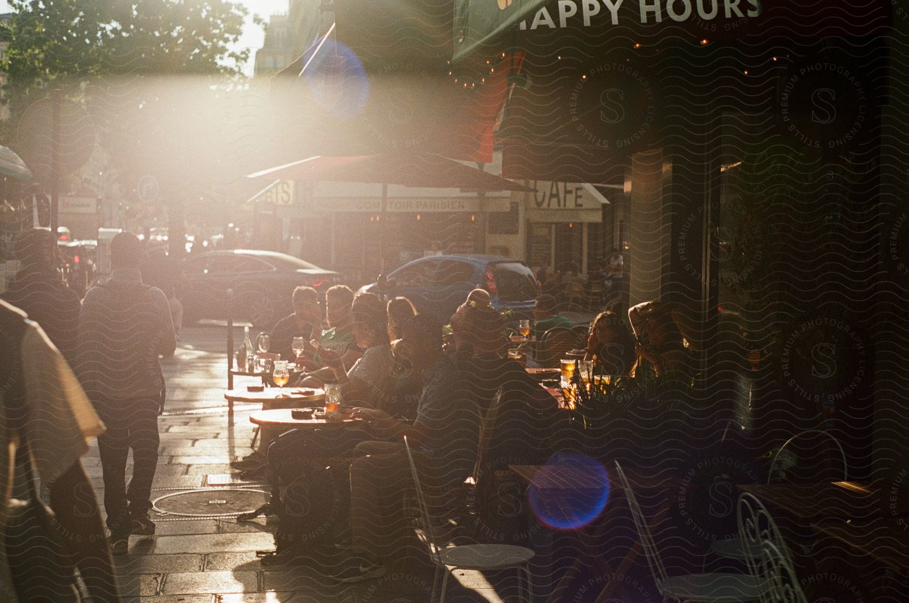 Several people sitting on chairs and tables on the street next to an establishment and people passing by behind