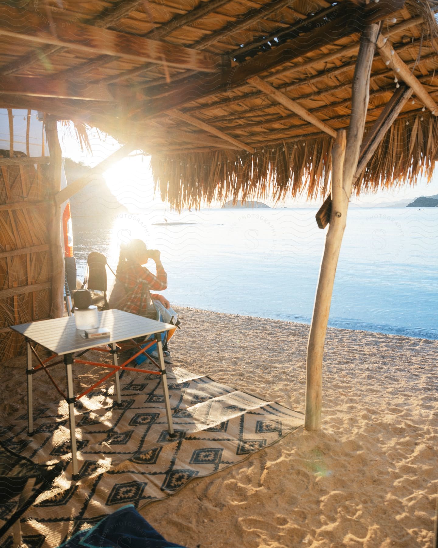 A person is sitting in a chair on the beach looking at the sea which is calm and there is a lack of sunlight illuminating the wooden frame that is above the person