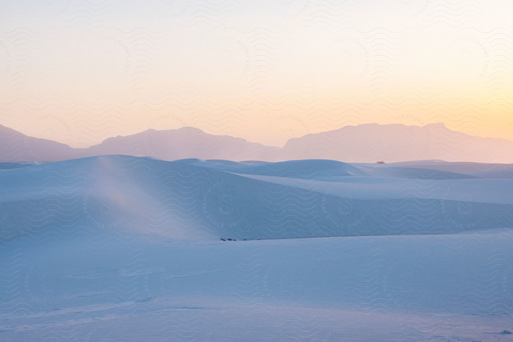 A serene landscape of smooth white sand dunes with distant mountains under a soft gradient sky during sunset