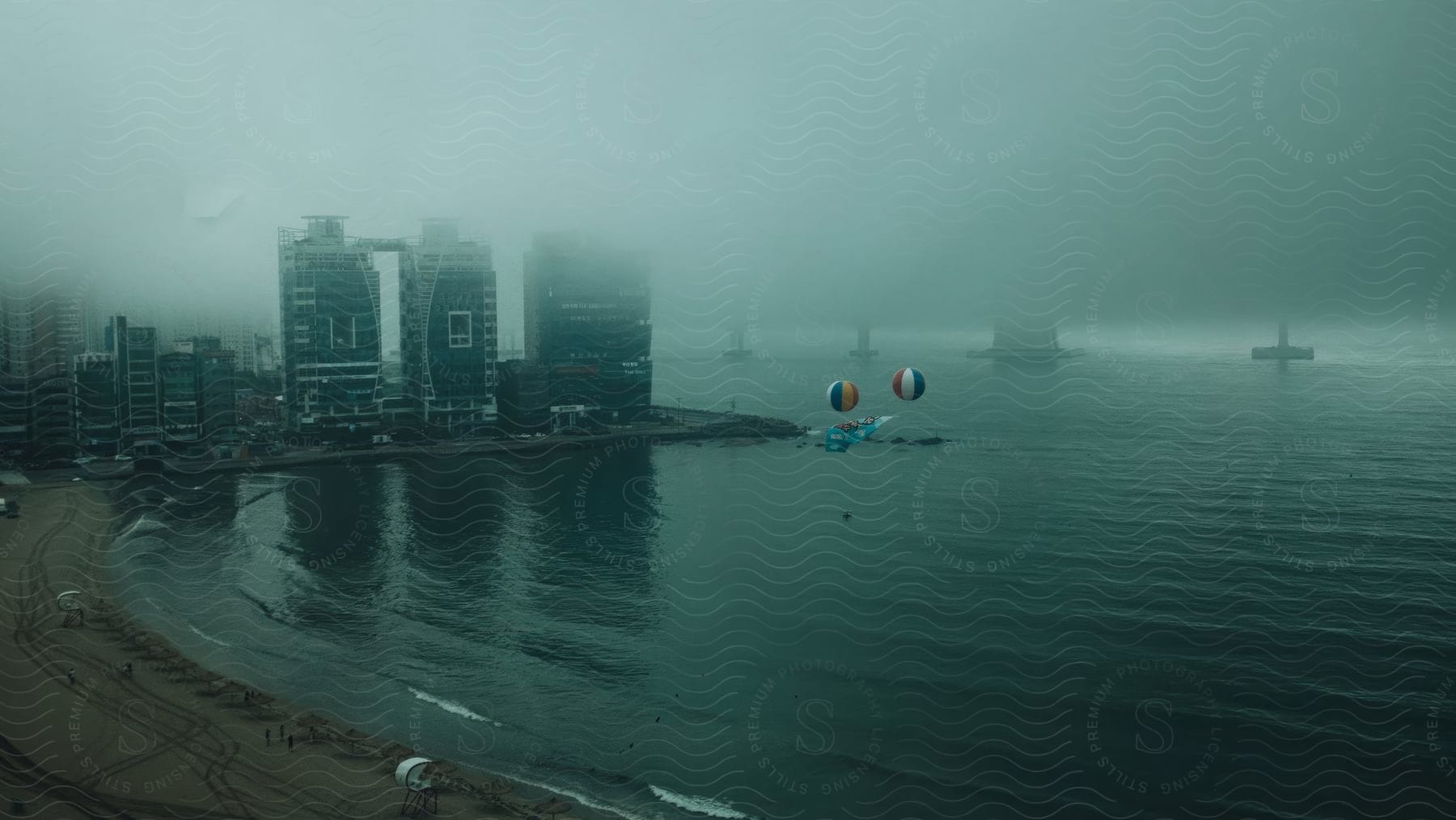 A beach with two balloons flying over in cloudy weather with a lot of fog