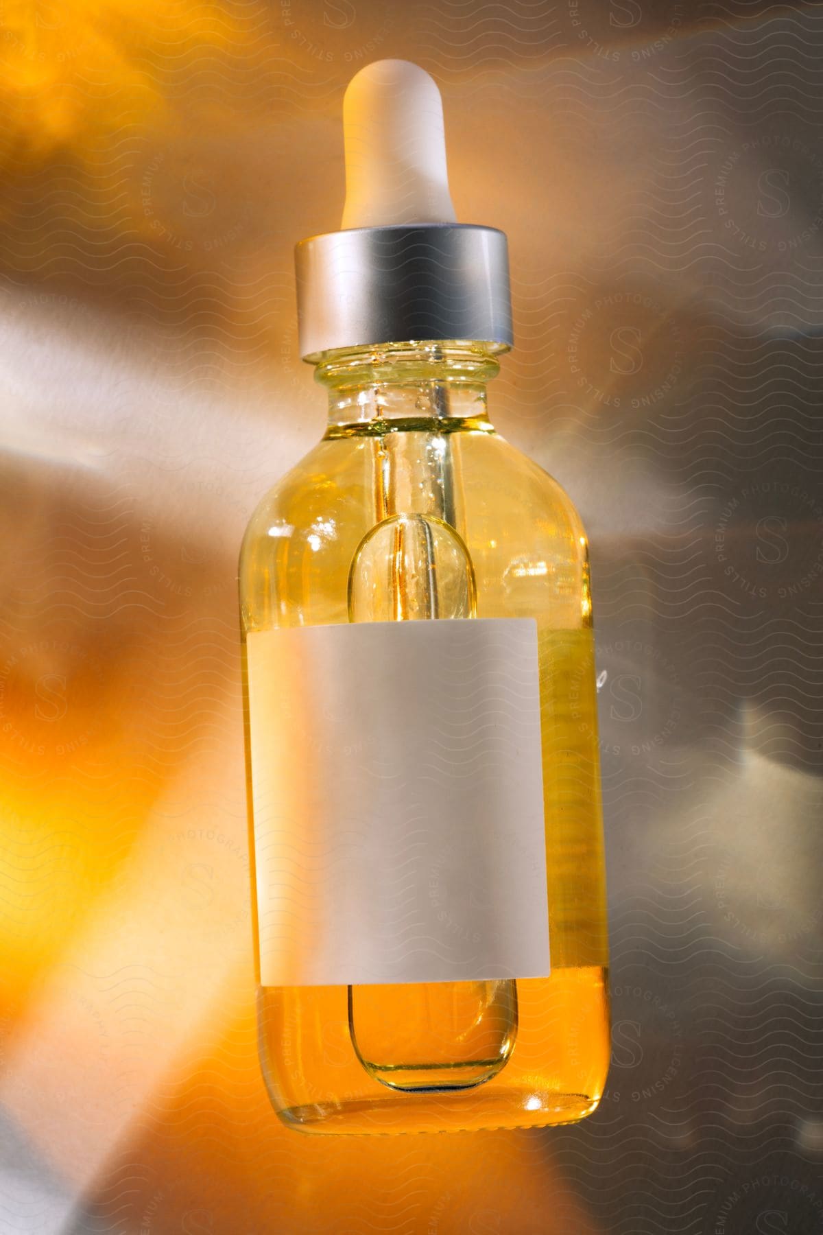 A transparent glass bottle with a yellow liquid, illuminated by a golden light. The bottle has a blank white label and a cap with a dropper.