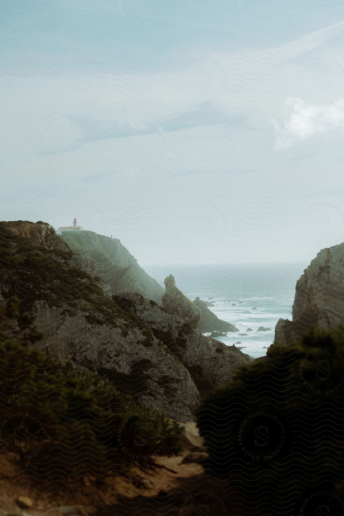 A serene coastal landscape with a lighthouse perched on the edge of a rocky cliff, surrounded by green vegetation and the ocean.